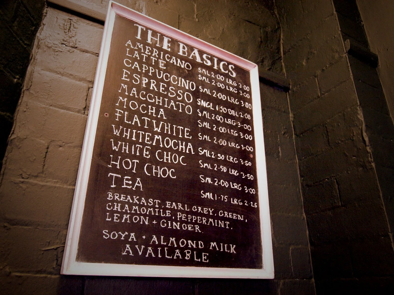 Menu board on brick wall listing beverages, including American, latte, cappuccino, espresso, macchiato, mocha, flat white, white mocha, white choc, hot choc, tea, and options for soy and almond milk.