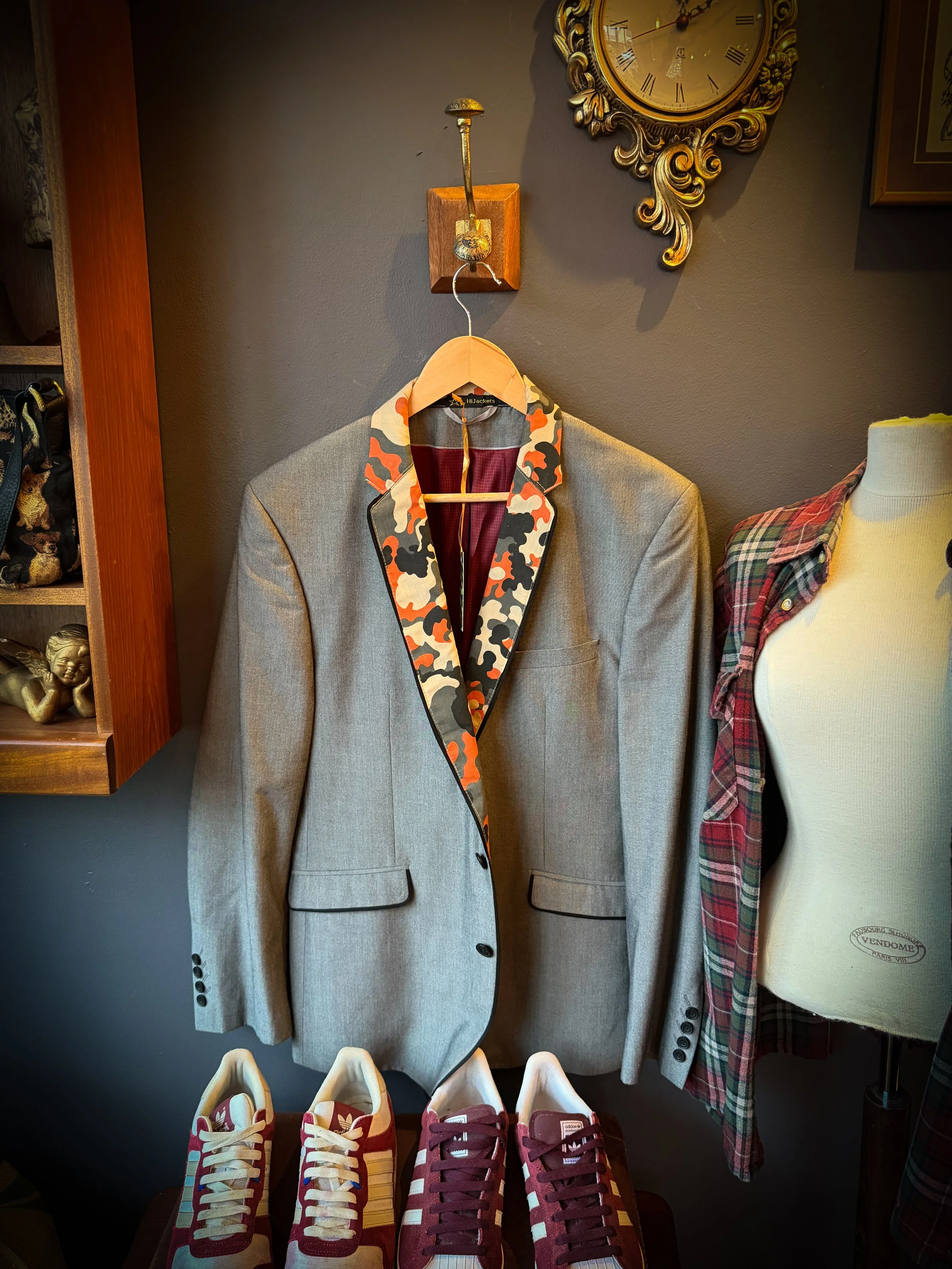 A gray blazer with a camouflage-patterned lapel hanging on a wooden hanger on a wall. Below the blazer are three pairs of sneakers in red, yellow, and burgundy. To the right, there is a mannequin torso dressed in a plaid shirt. The background feature