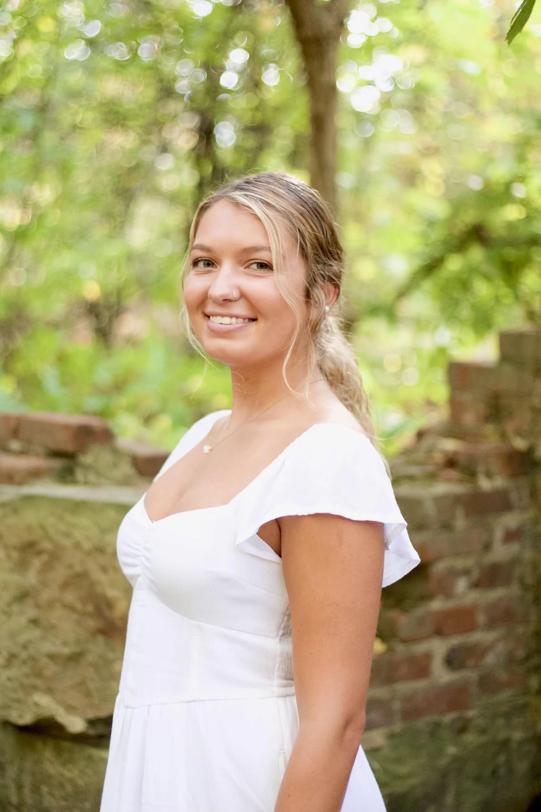 A young woman with blonde hair tied in a braid, smiling outdoors against a background of green leaves and trees, wearing a white off-shoulder dress.