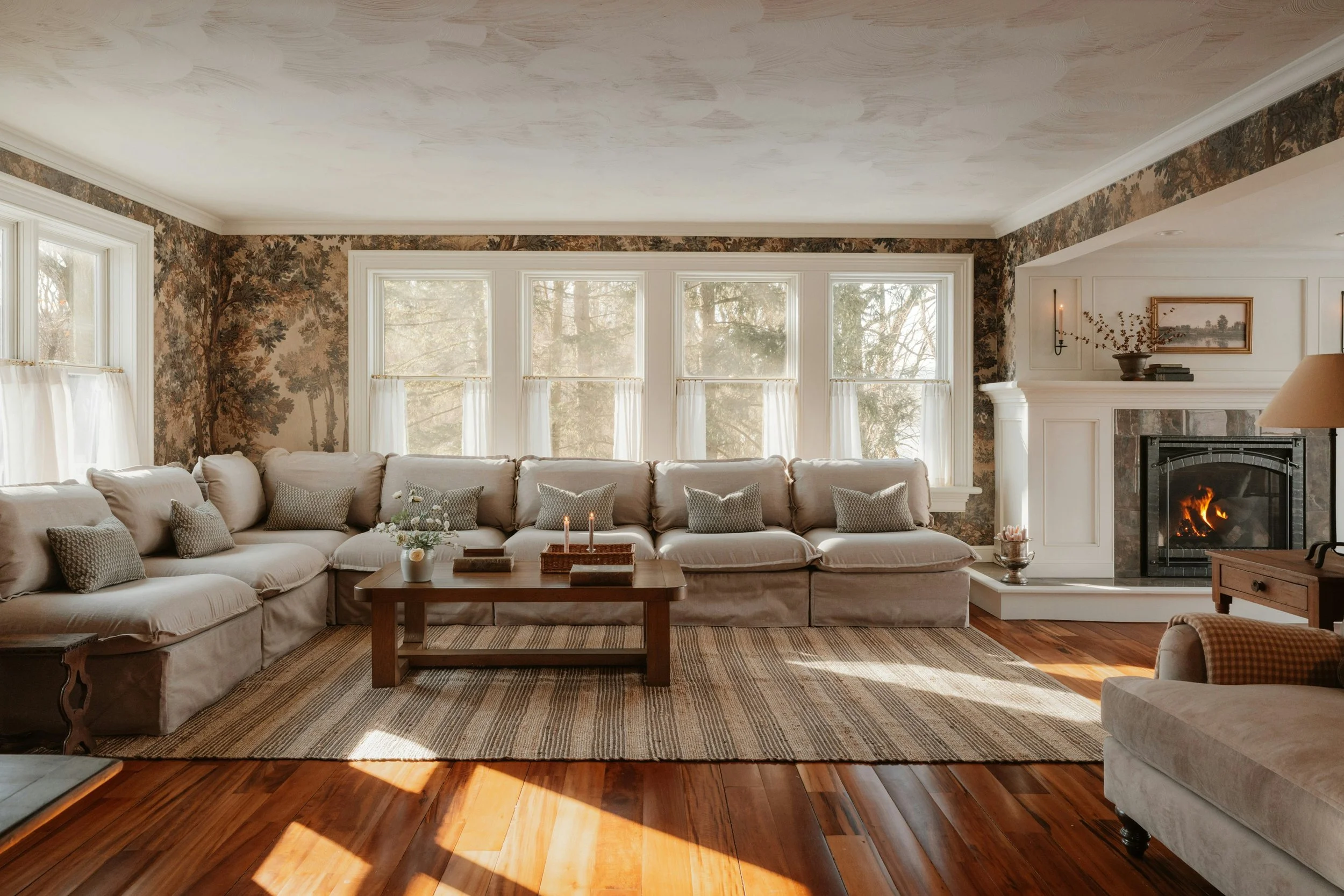 Living room with beige sectional sofa, wooden coffee table, fireplace, large windows with white curtains, floral wallpaper, and hardwood floors.