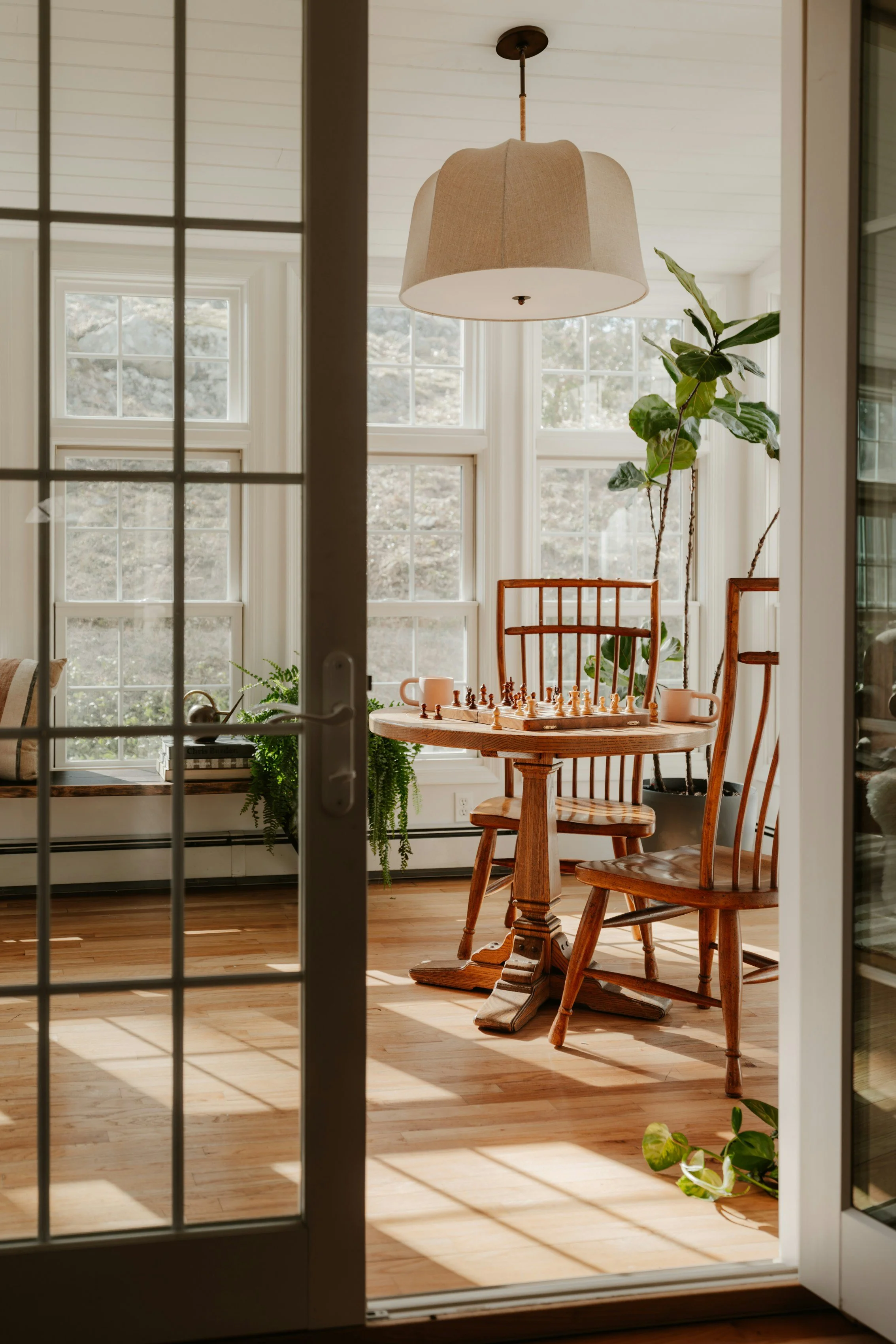 Sunlit room with large windows, a round wooden table with chess set and cups, wooden chairs, a hanging lampshade, and potted plants, seen through a glass door with grid pattern.