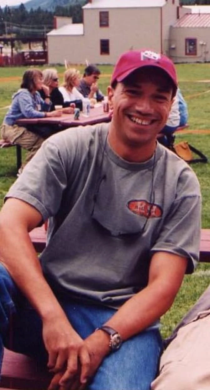 A smiling man wearing a red cap and a gray T-shirt, sitting outdoors at a picnic table with a group of people in the background.
