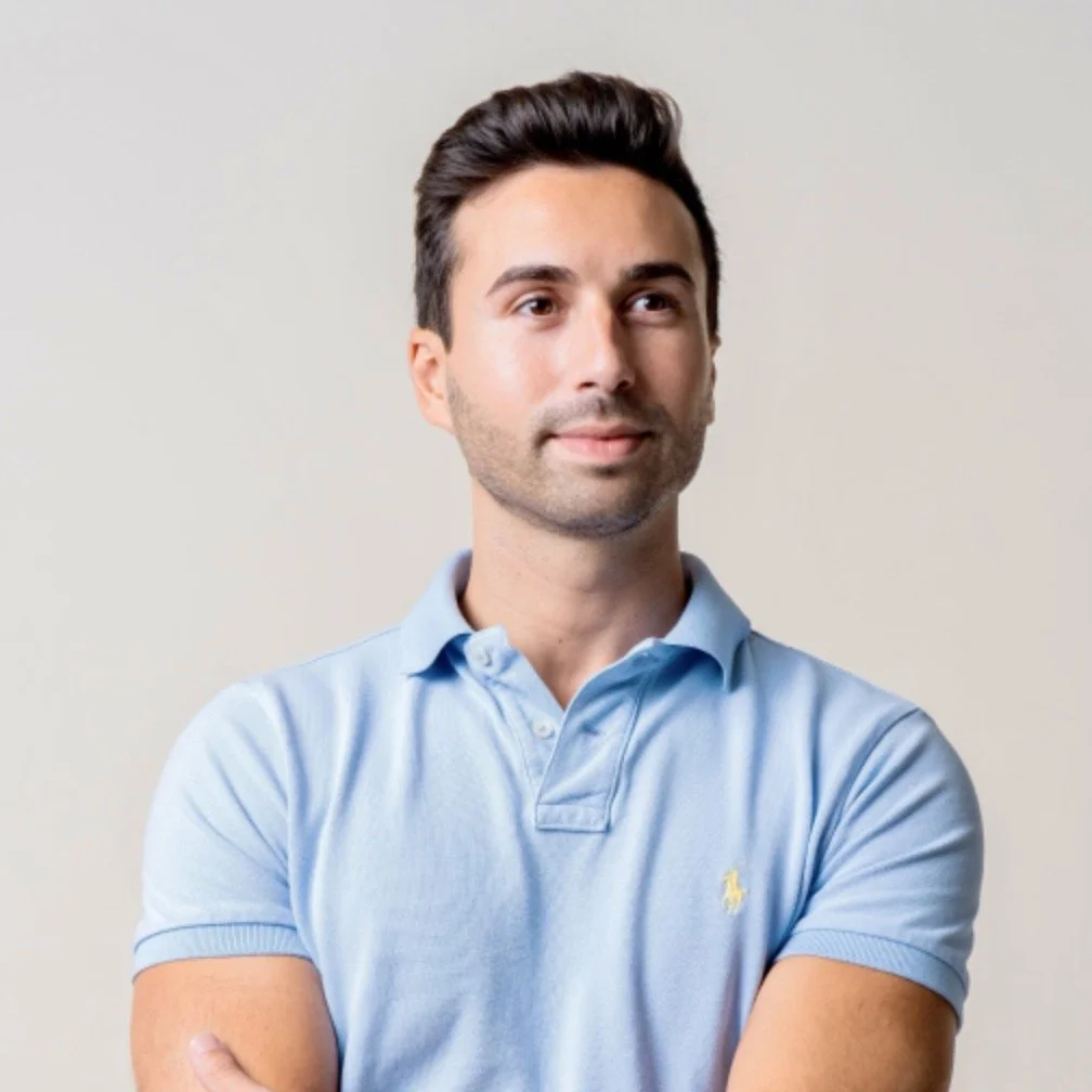 A man with dark hair and light skin, wearing a light blue polo shirt, standing with arms crossed against a plain light background.
