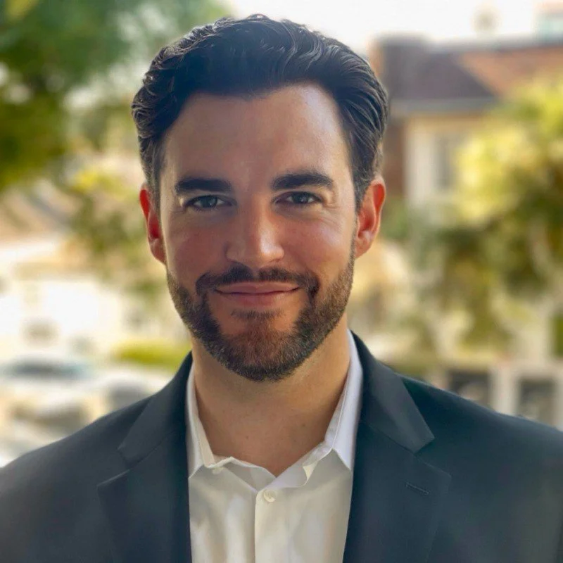 A man with dark hair and a beard in a suit outside with trees and houses in the background.