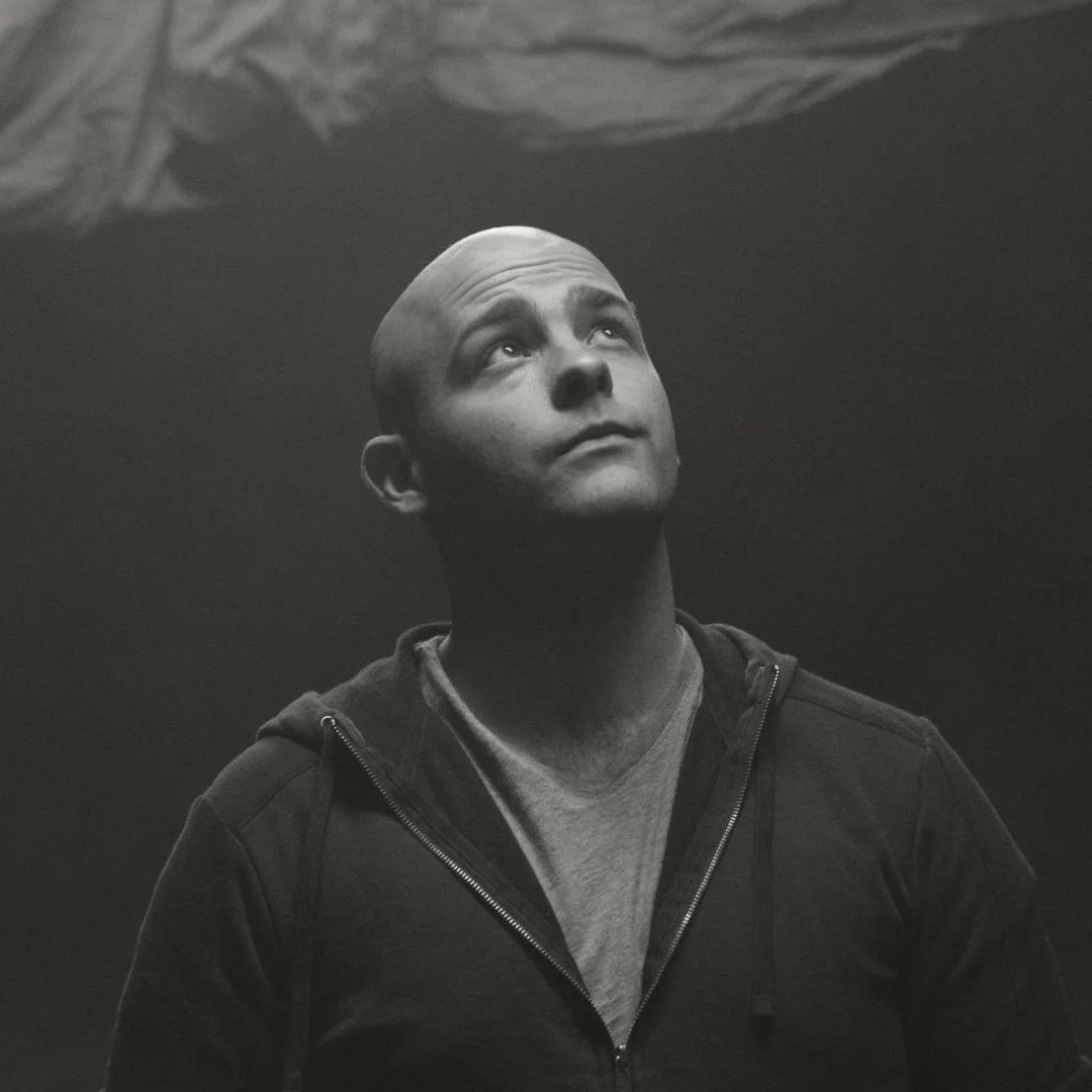 A black and white photo of a man with a shaved head looking upward. He is wearing a hoodie and a T-shirt indoors.