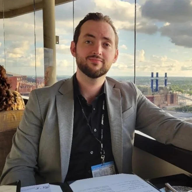 A man with dark hair and a beard, wearing a gray blazer and black shirt, sitting at a table with a cityscape view through large windows behind him.