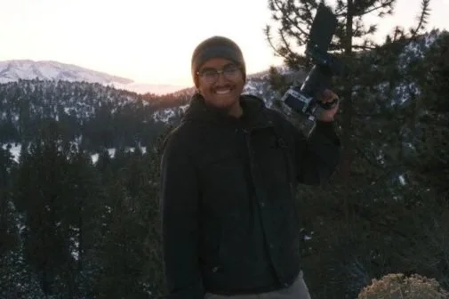 A man with glasses smiling while holding a camera in a mountainous forest at sunset.