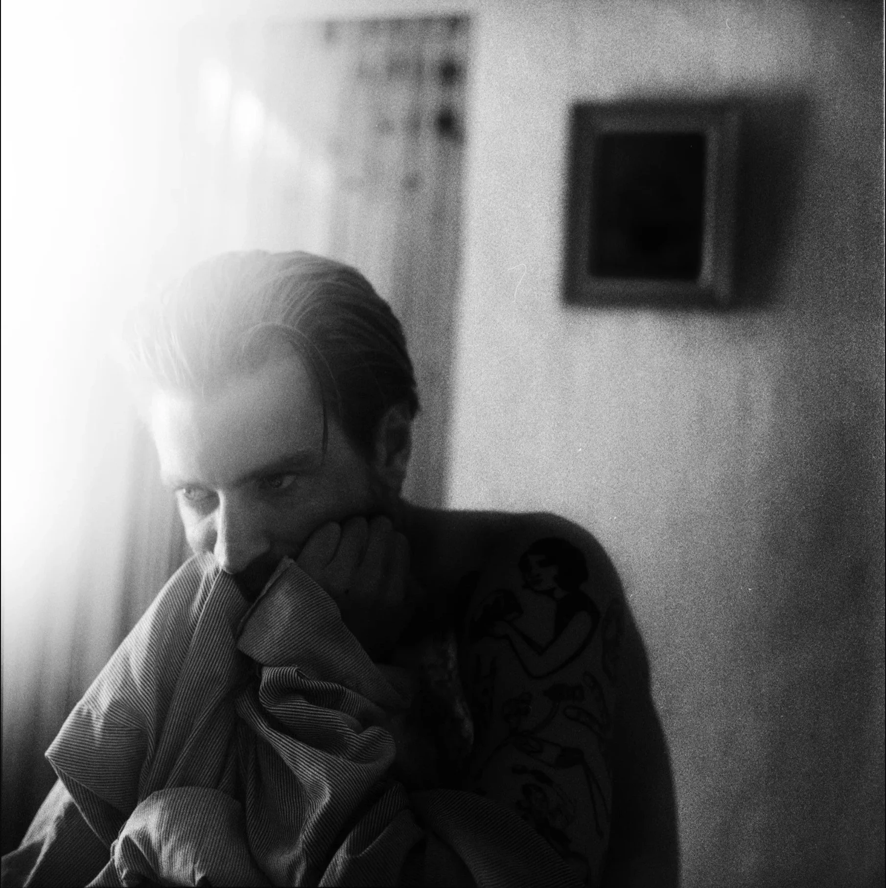 Black and white photo of a person with light hair, partially covering their face with their hand, looking thoughtfully to the side, with an arm tattoo visible. Light source from the left side, with a picture on the wall in the background.