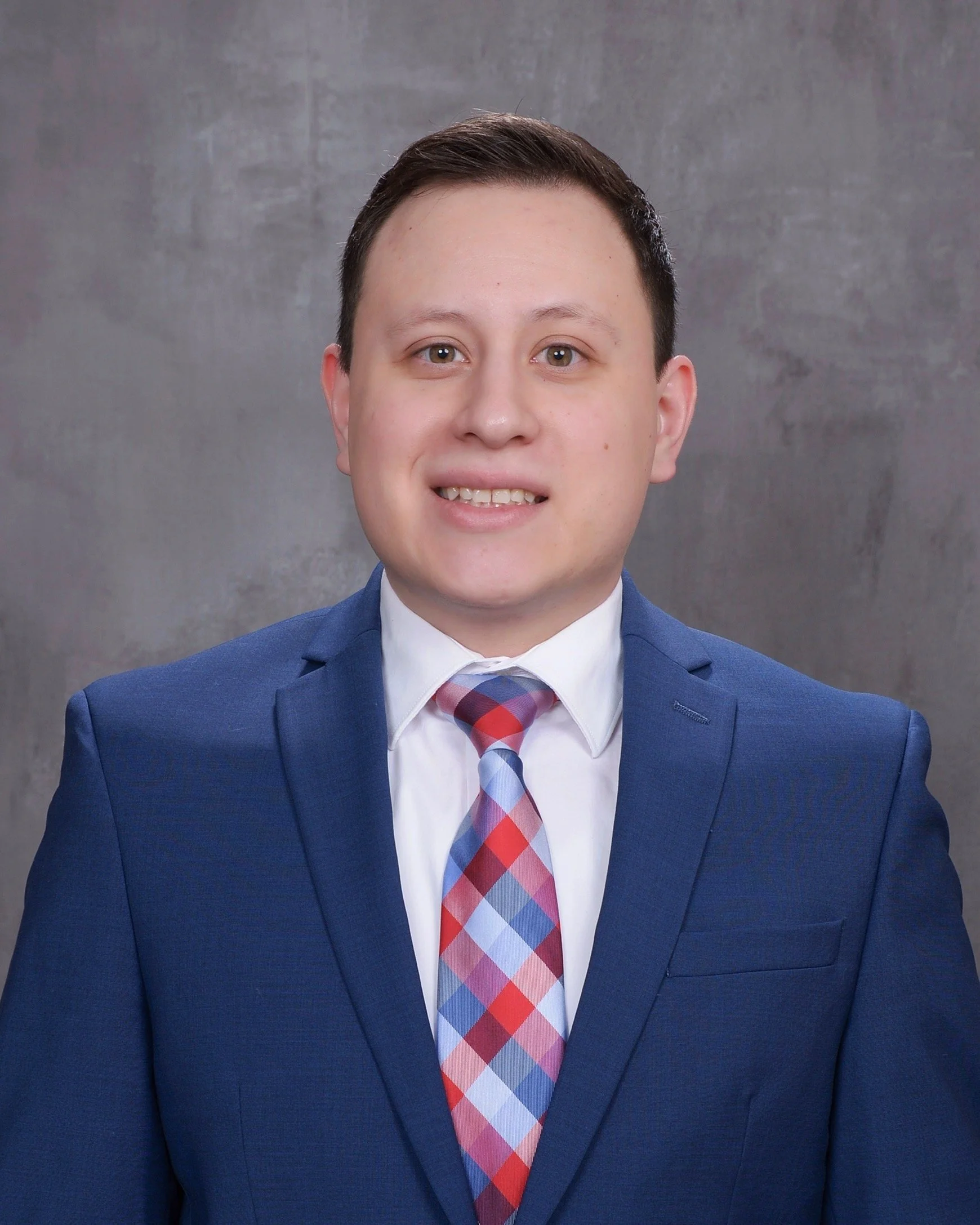 A young man in a blue suit, white shirt, and a colorful plaid tie, smiling in front of a gray background.