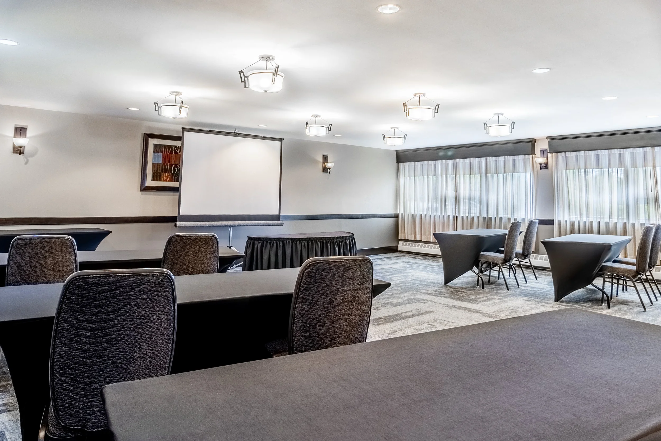 A conference room with several tables and chairs, a projection screen, and multiple ceiling lights, with large windows and curtains at Best Western Plus Chocolate Lake.