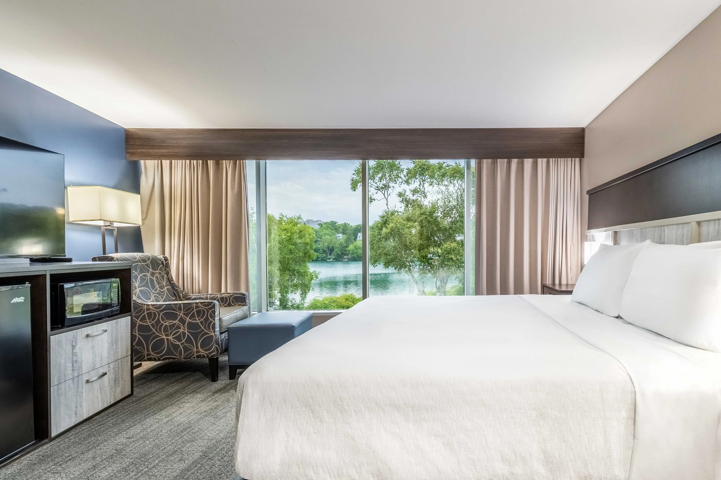 A Best Western Plus Chocolate Lake Hotel room with a large window overlooking a lake with trees, a bed with white linens, a patterned armchair, a small microwave and mini fridge, and a TV on a stand.