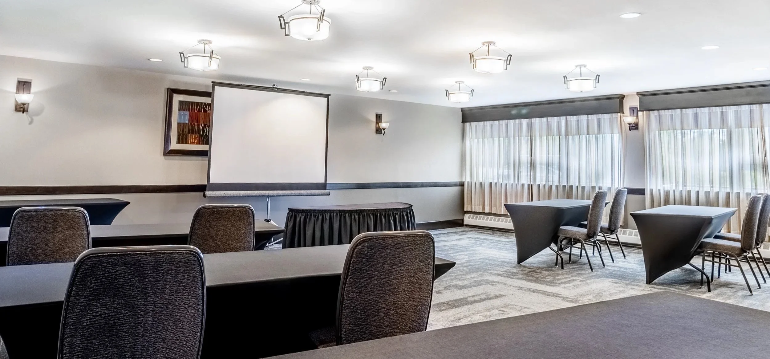 A conference room with tables, chairs, a projection screen, and large windows with curtains at Best Western Plus Chocolate Lake.