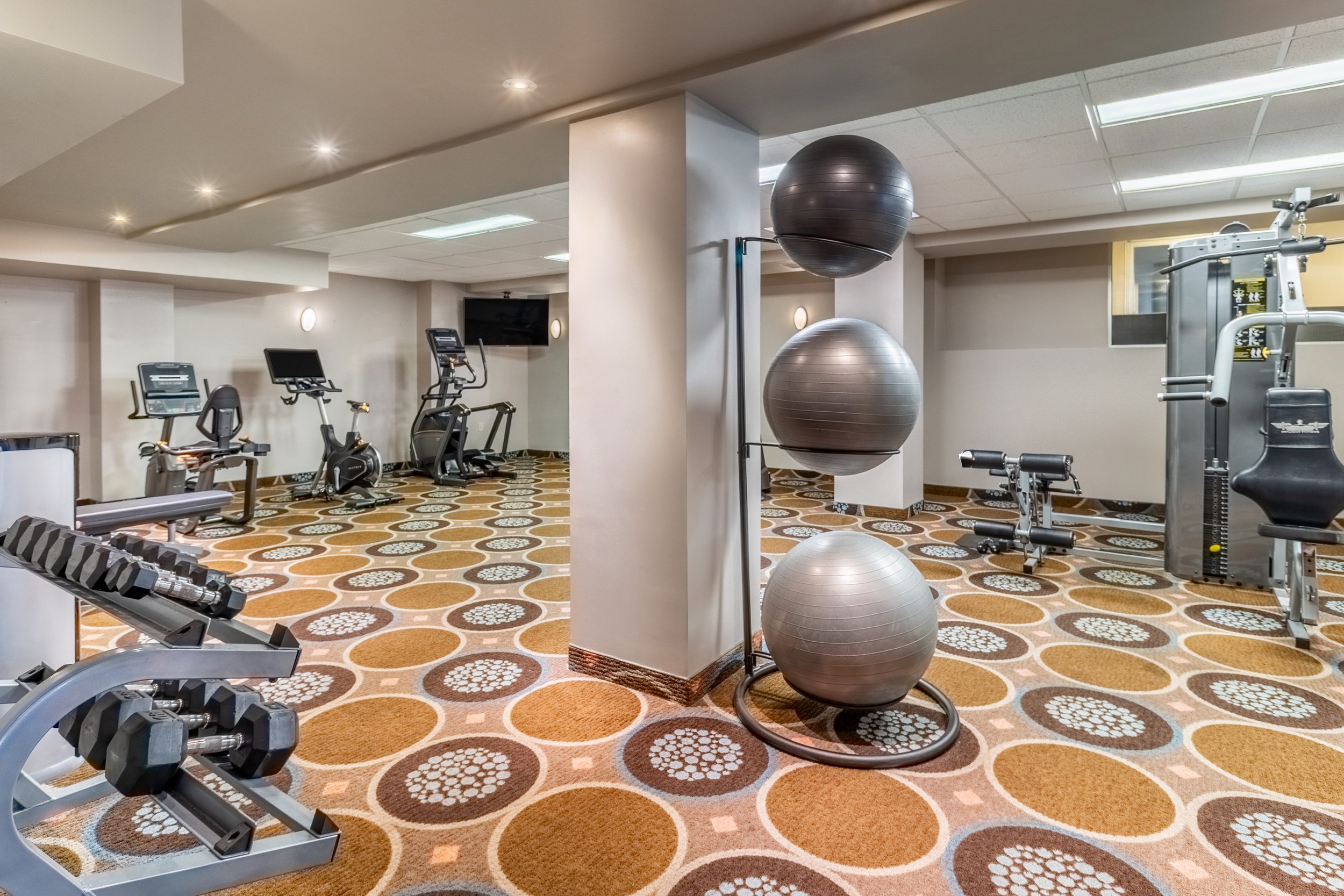 Indoor gym at Best Western Plus Chocolate Lake, with exercise equipment including treadmills, ellipticals, dumbbells, and stability balls on a patterned carpet, with ceiling lights and a wall-mounted TV.