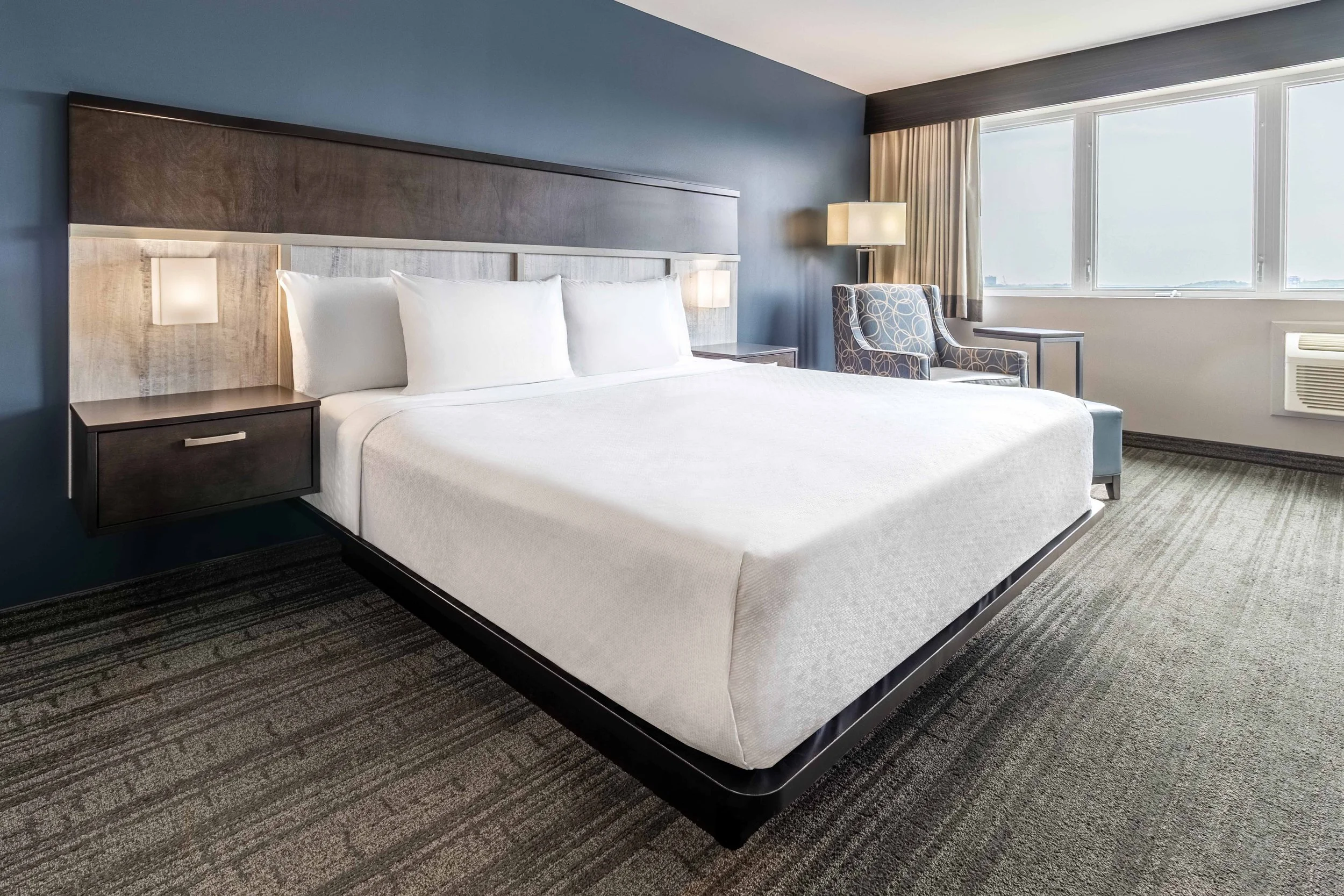 A Best Western Plus Chocolate Lake hotel room with a large bed, white linens, a headboard with built-in lights, a nightstand, an armchair with a patterned fabric, a floor lamp, and large windows with curtains showing a view outside.