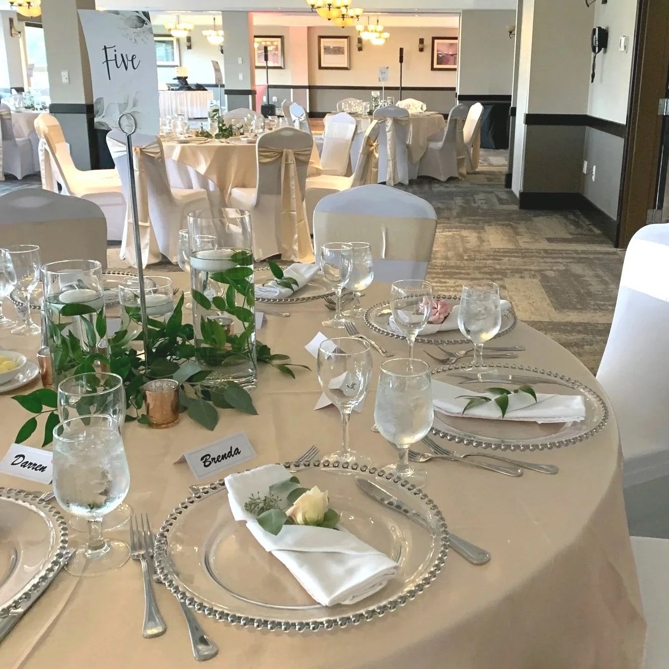 Wedding reception table, in one of Best Western Plus Chocolate Lake's ball rooms, decorated with greenery, candles, white napkins, and place cards with names, surrounded by elegant chairs and additional tables in the background.