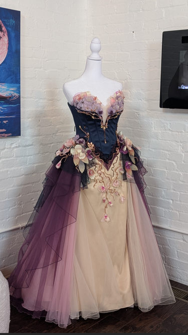 A mannequin dressed in a strapless gown with a beige skirt and purple tulle overlay, adorned with floral appliqués and gold embroidery, displayed against a white brick wall.