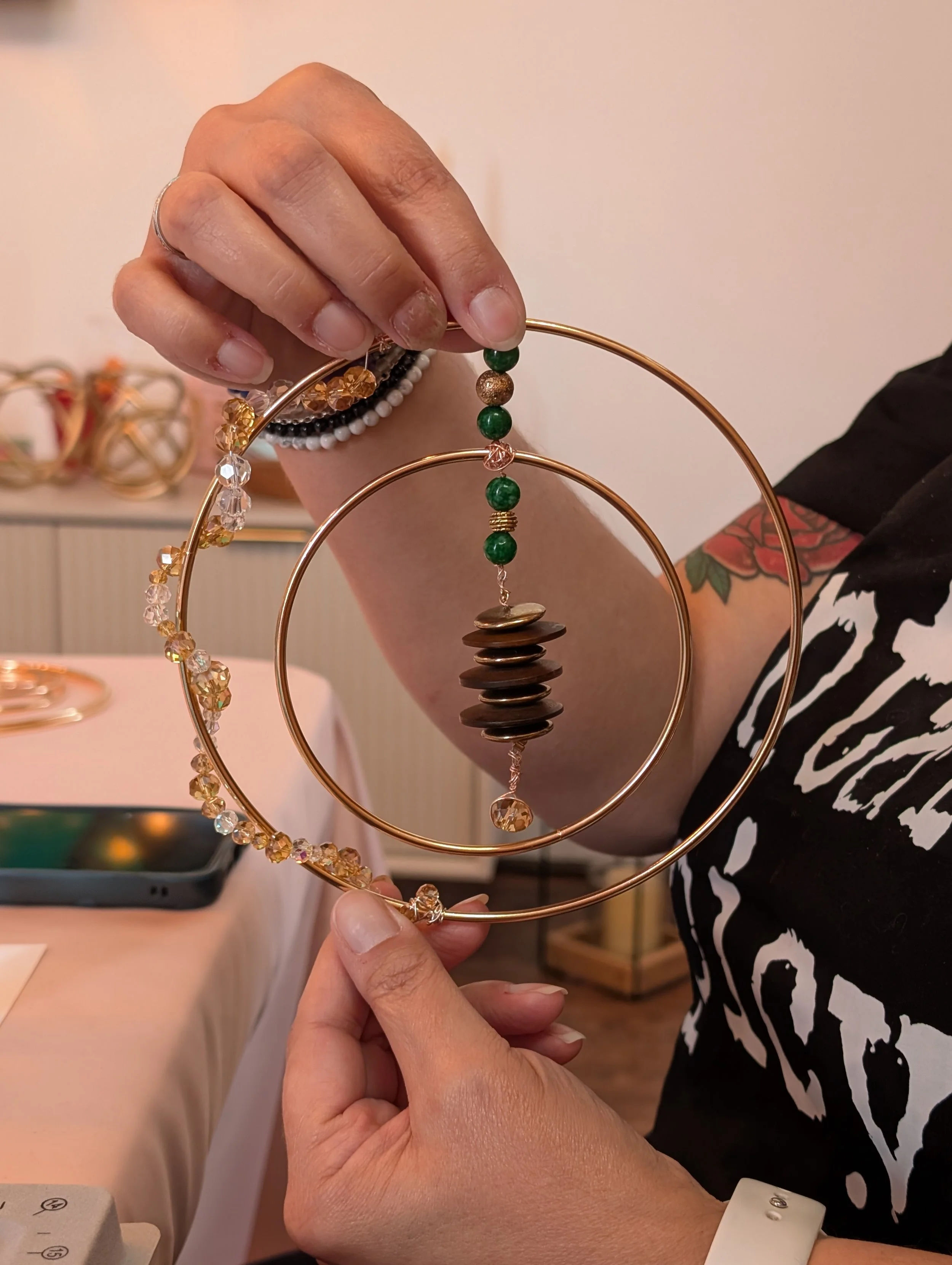 Person holding a gold circular jewelry display stand with a beaded bracelet and a string of green and pink beads, with another person's hand in the background, a tattooed arm, and part of a black and white patterned shirt.