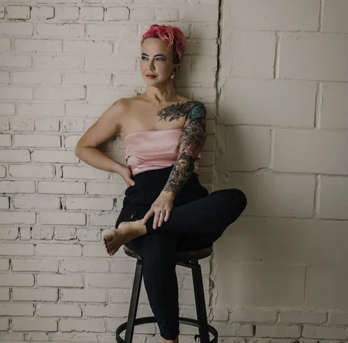Shannon Elizabeth, with short pink hair and a floral tattooed arm, seated against a white brick wall