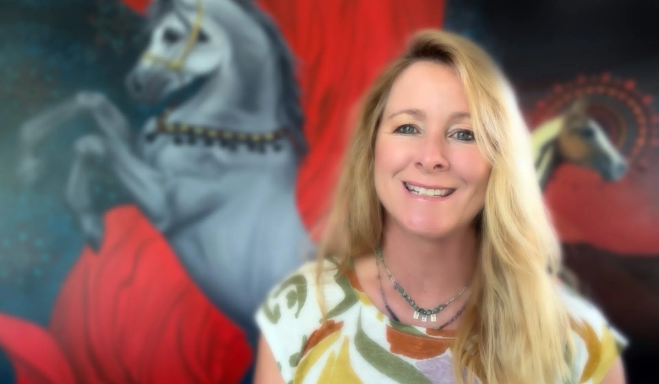 A smiling woman with long blonde hair wearing a patterned shirt, standing in front of a colorful background featuring abstract art with a tiger and a geometric design.