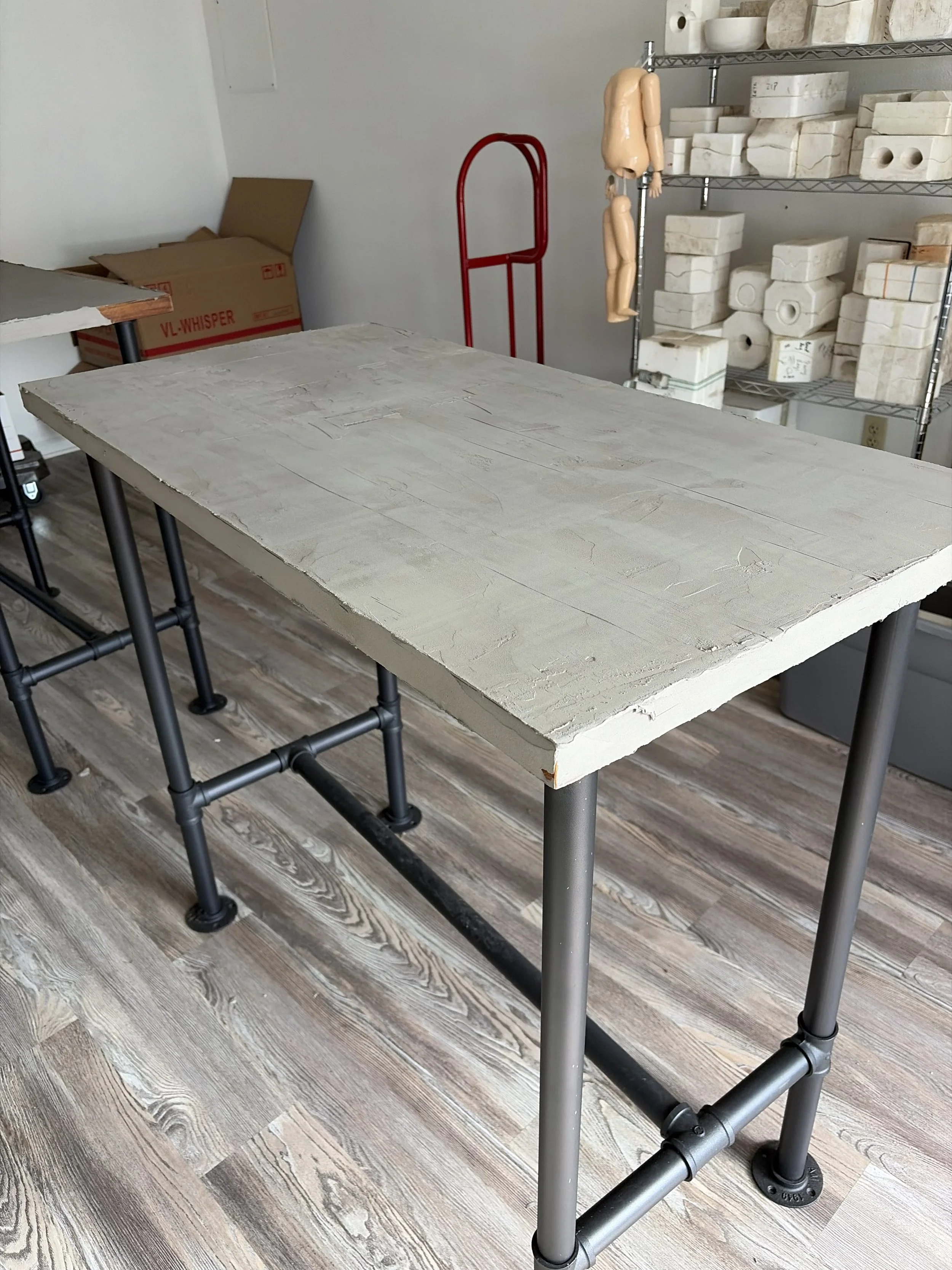 A metal table with a concrete top in a workshop or studio. In the background, there are shelves with blocks of marble or stone, a red dolly, and a mannequin torso. The floor is wood with a weathered appearance.