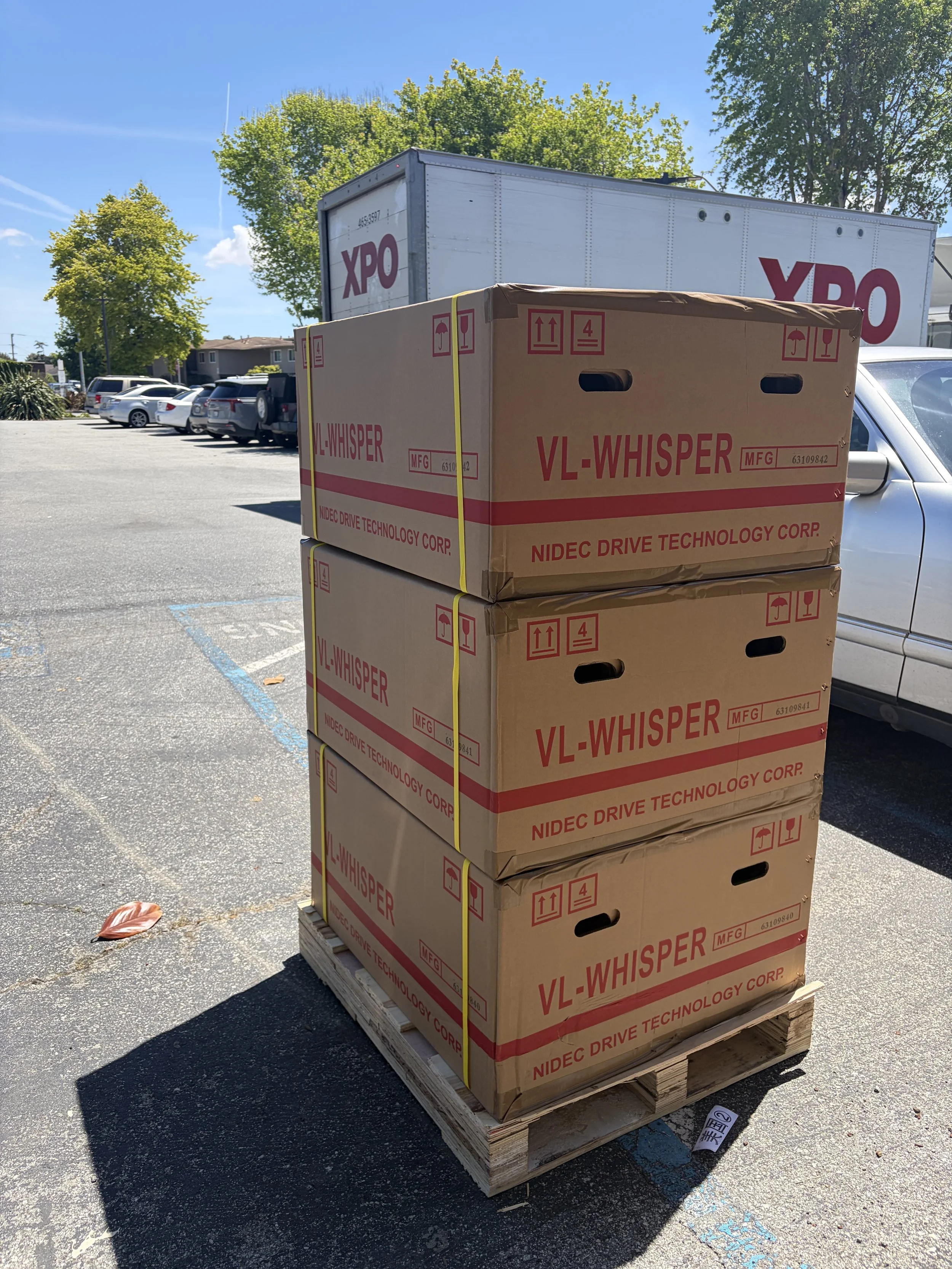 Four stacked cardboard boxes labeled VL-WHISPER on a wooden pallet in a parking lot, with cars, trees, and a building with a direct-to-consumer truck in the background.