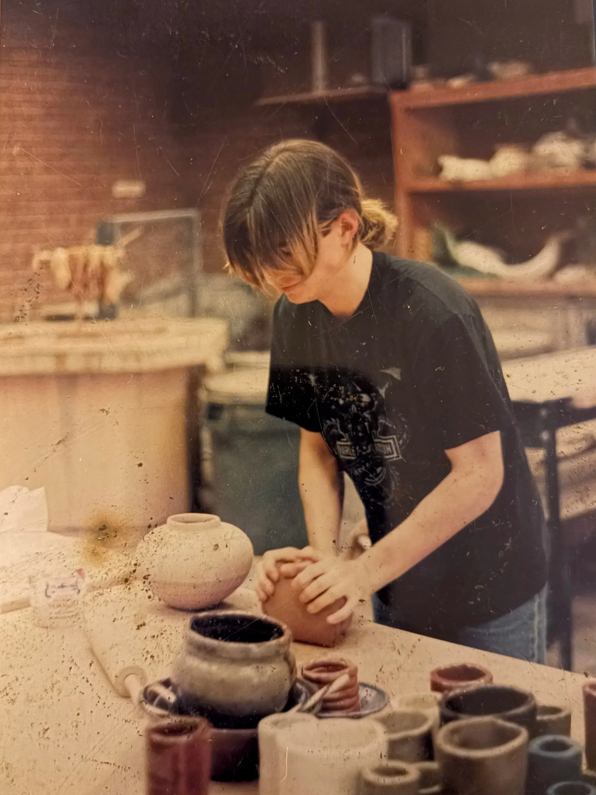 A person with shoulder-length hair, wearing a black t-shirt, working on a pottery wheel in a pottery studio. Various pottery tools and finished pottery pieces are on the work surface in front of them.