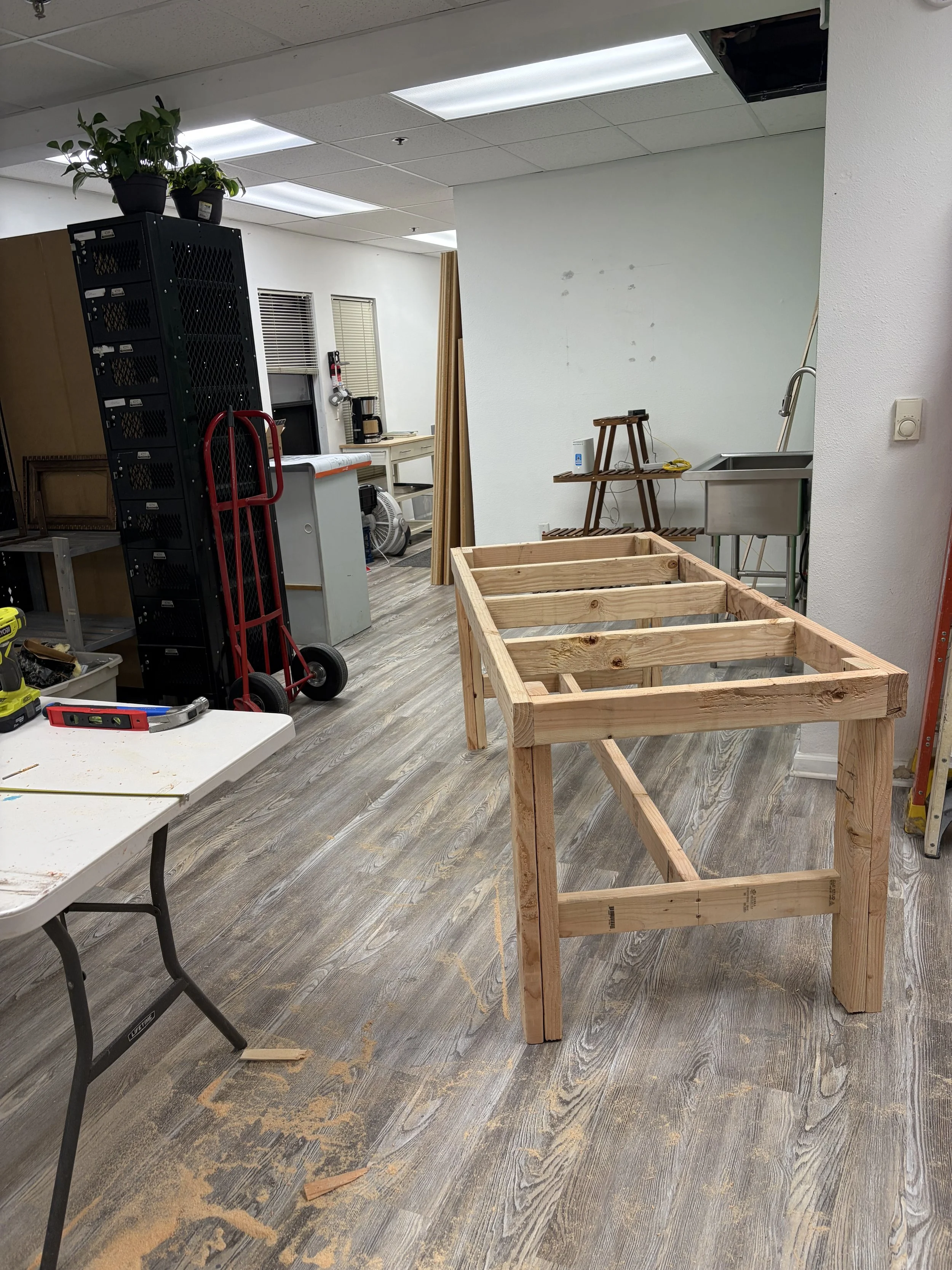A partially constructed wooden frame on a hardwood floor in an indoor space, with various tools, a hand truck, and supplies scattered around the room, and some wall and ceiling damage visible.