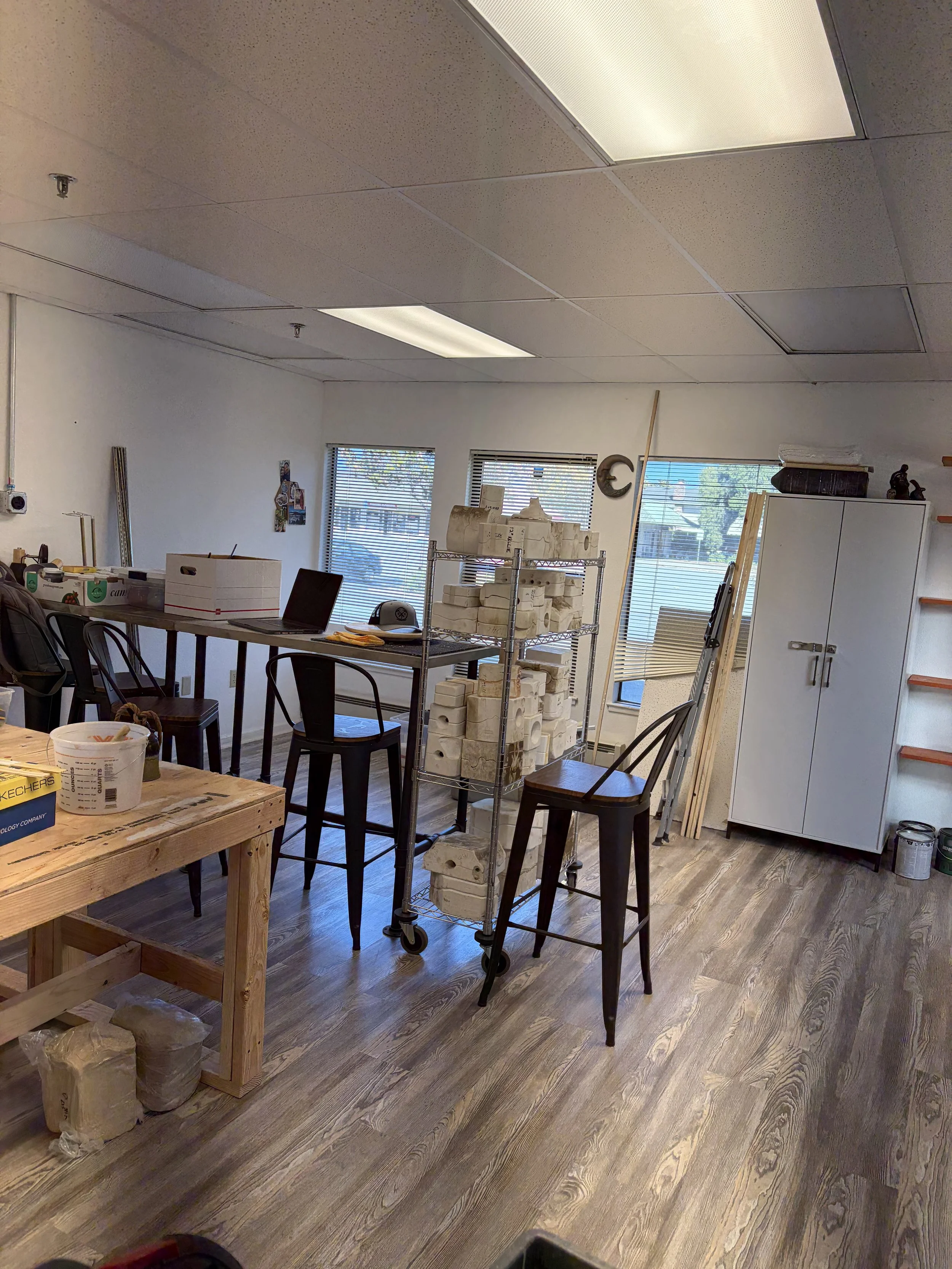An office or workshop with a wooden table, black chairs, a metal cart filled with white bricks, and a white cabinet. There are windows with blinds, some tools and objects on shelves, a laptop on the table, and a flooring that looks like wood.