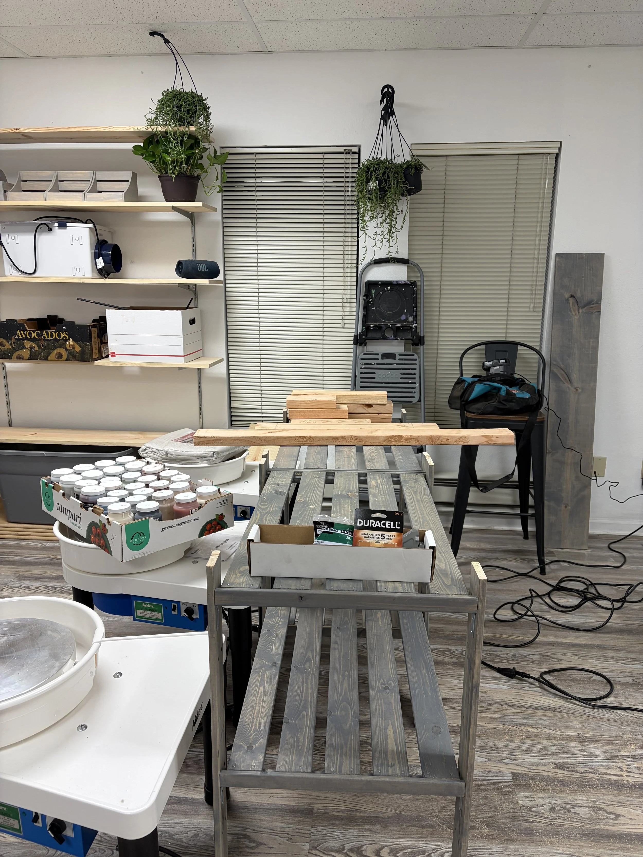 Workshop with tables, shelves, plants, and various tools and supplies, including jars, a saw, and a box of batteries.