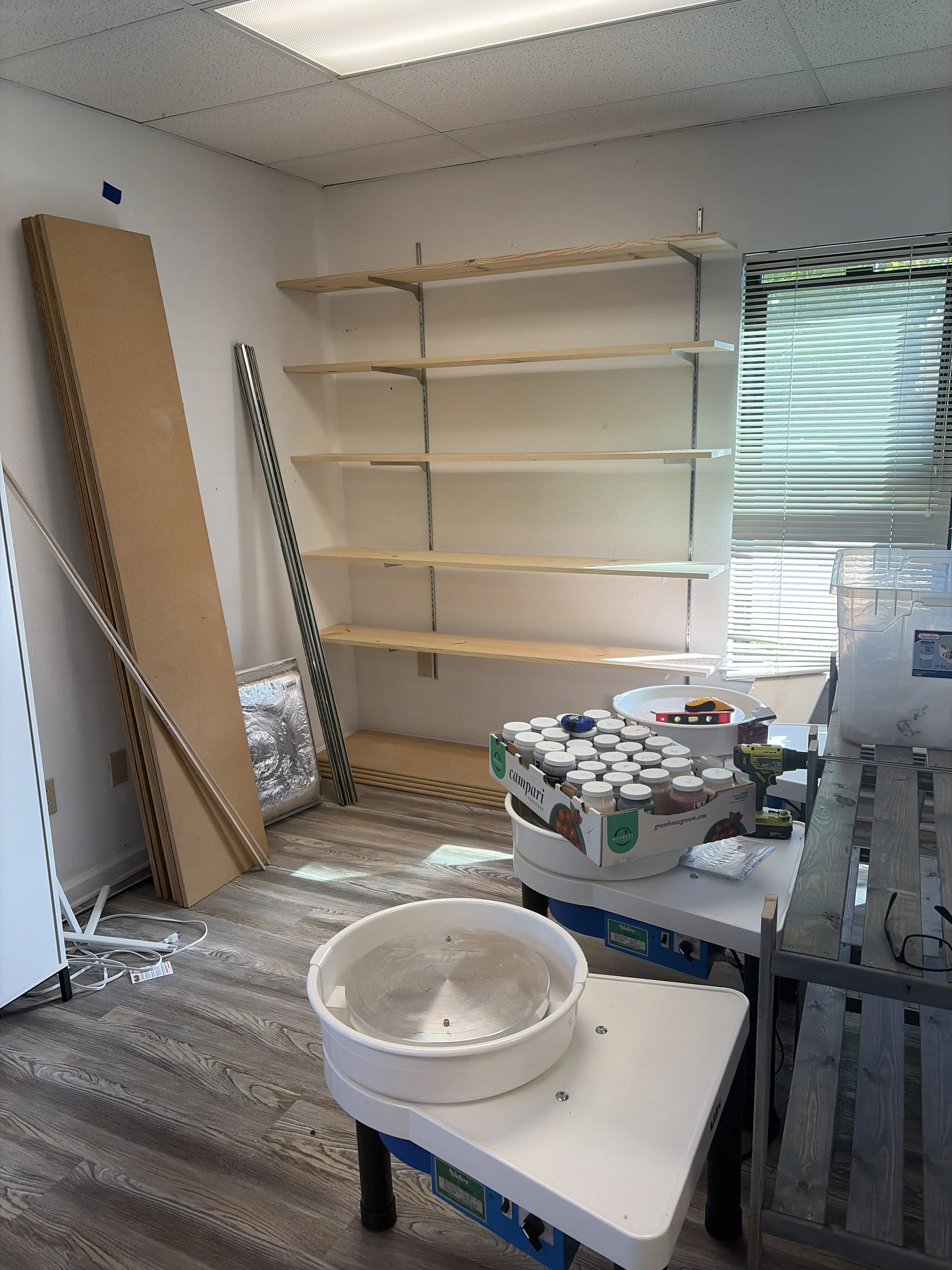 Room with empty wooden shelves on wall, various tools, a large bucket and containers of paint or chemicals on tables, and some hanging hardware or metal bars leaning against the wall.