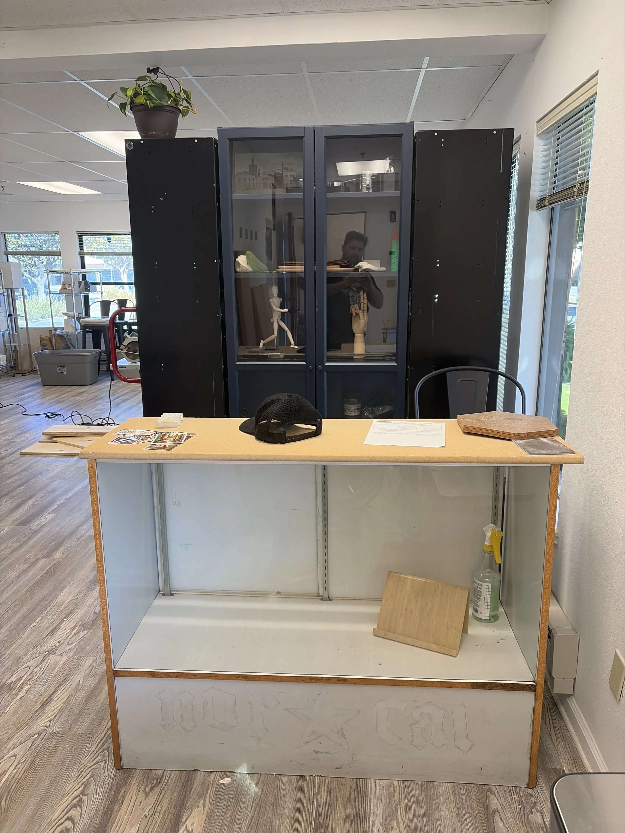 Empty display case with a wooden top in a store or workspace, with a spray bottle and wooden board on the floor beside it, and a black mesh chair in front.