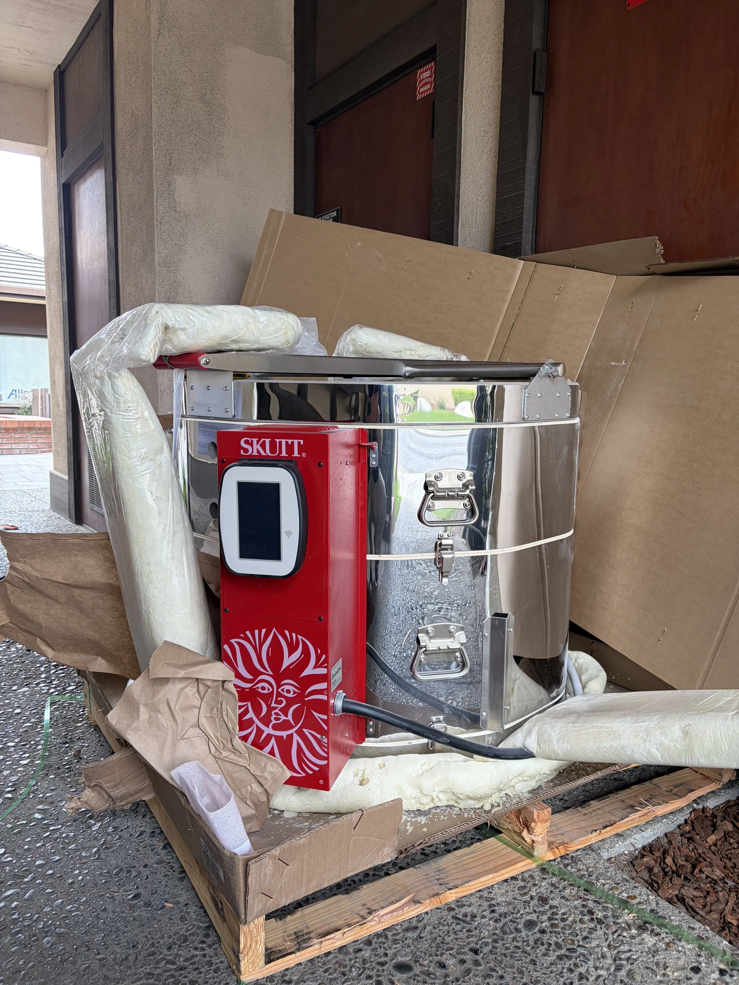 A Skutt KMT-1222 Electric Kiln, placed on a wooden pallet surrounded by cardboard and foam packaging. The machine has a small logo of a sun with the word 'SKUTT' on the red panel.