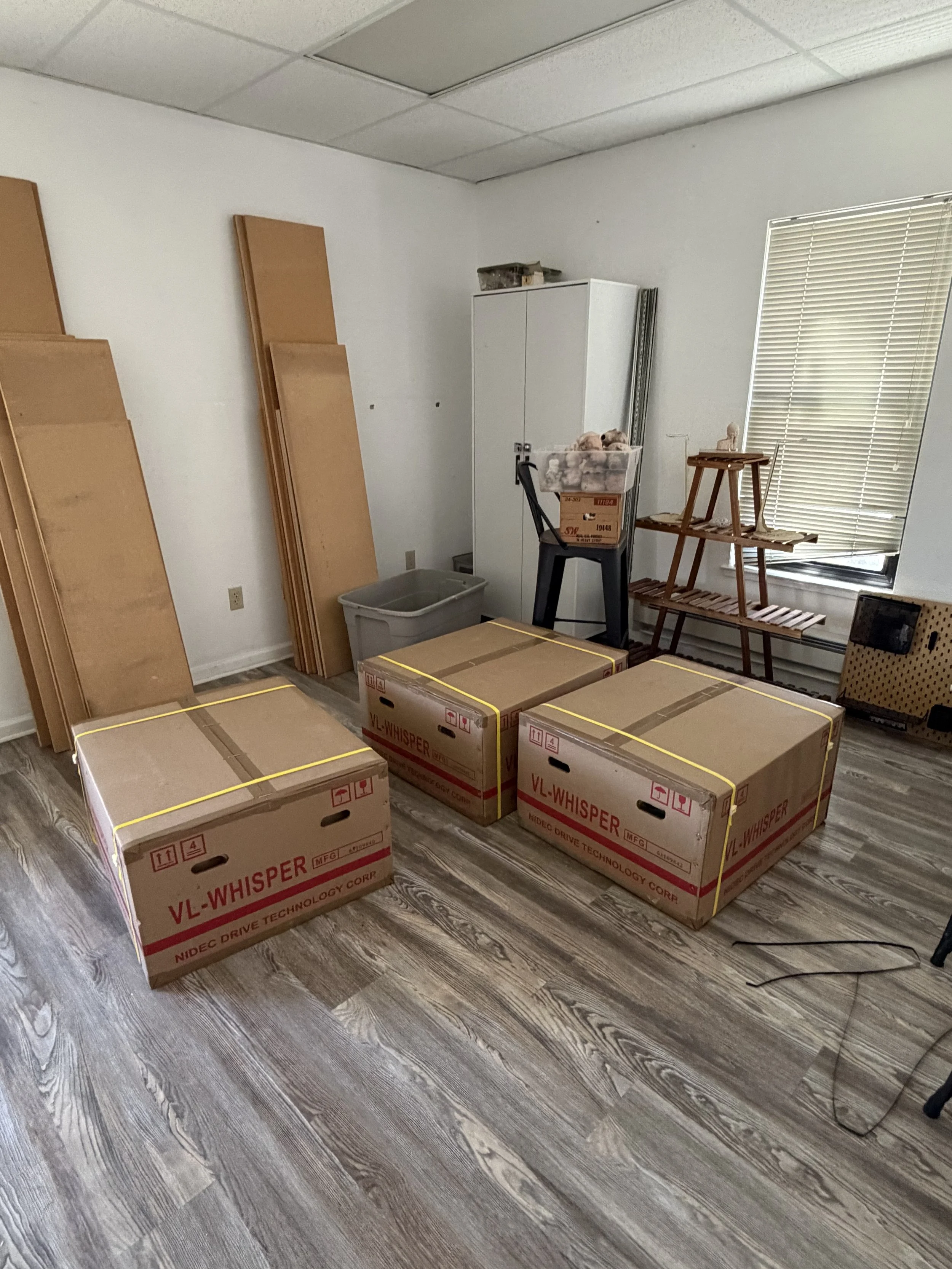 Room with boxes, shelving, window with blinds, cardboard boxes of Shimpo Electric Pottery Wheels labeled "VL-WHISPER" on wood floor.
