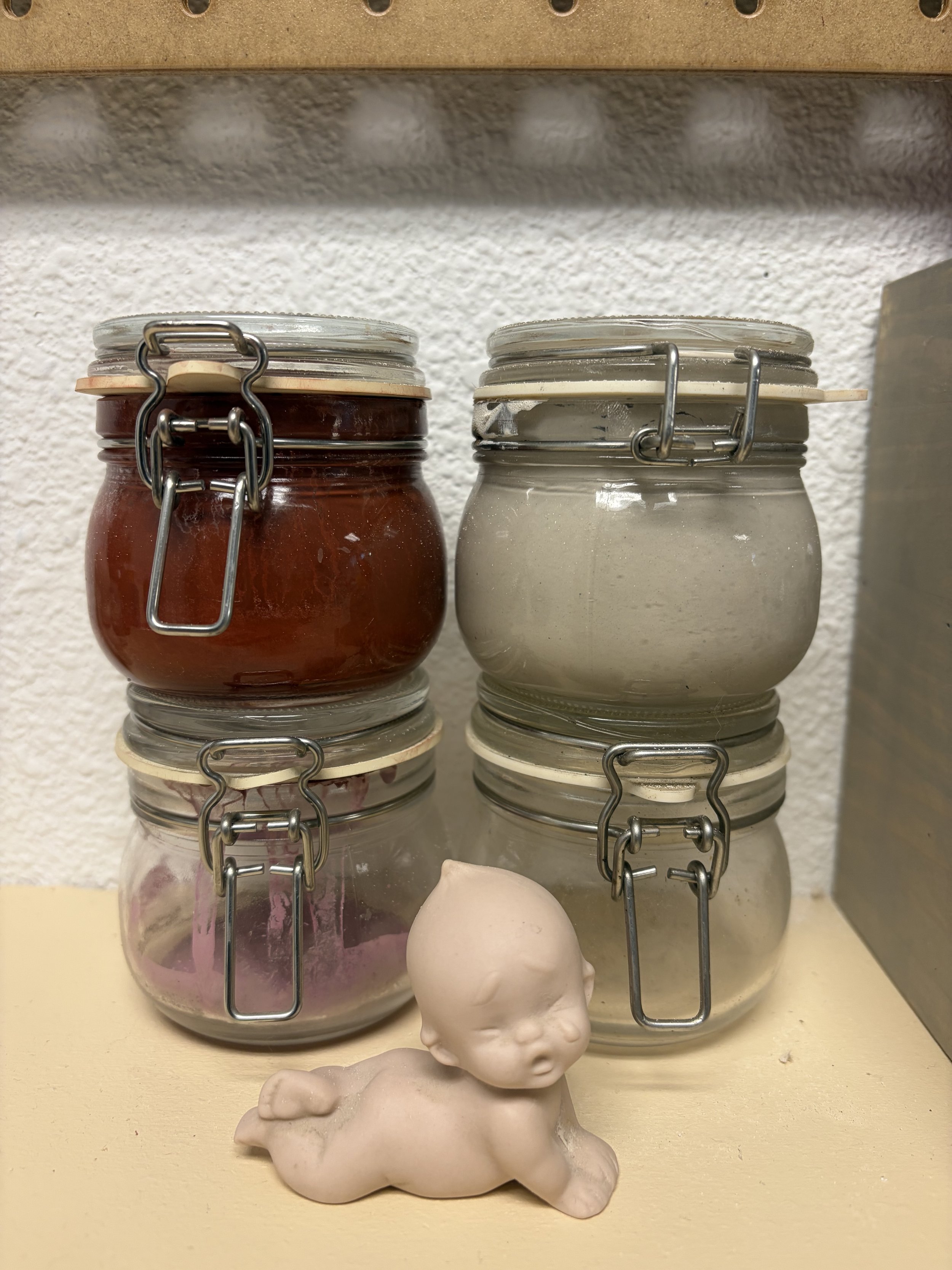 Six glass jars with metal clamp lids, containing various colored pastes or creams, are stacked two high on a shelf. A small white ceramic figurine of a sleeping dog is placed in front of the jars, against a textured white wall background.