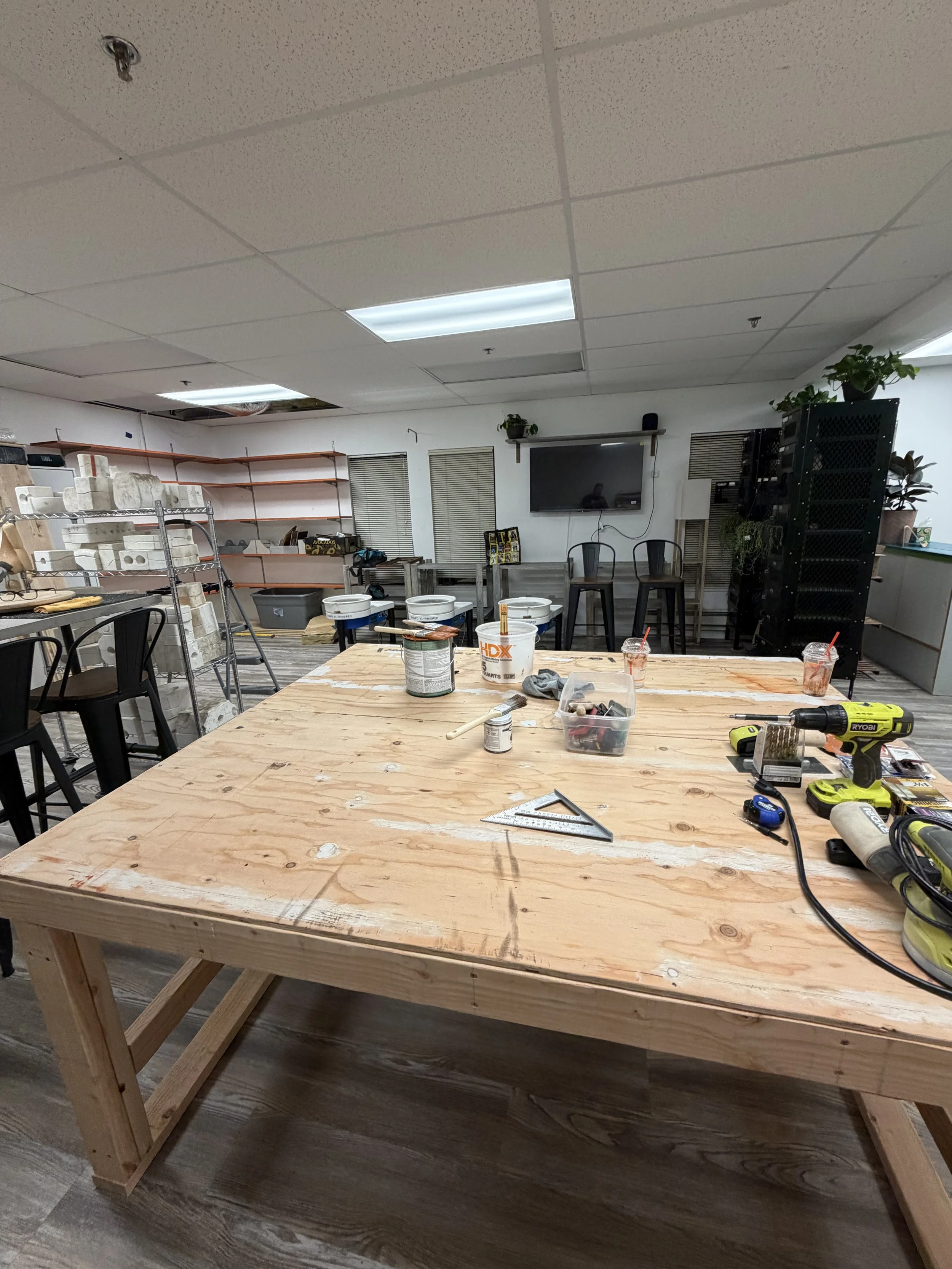 A study or workshop room with a large wooden worktable in the foreground, covered with tools, paint buckets, and cups with straws. In the background, there are shelves with materials, a TV on the wall, and various chairs and plants.