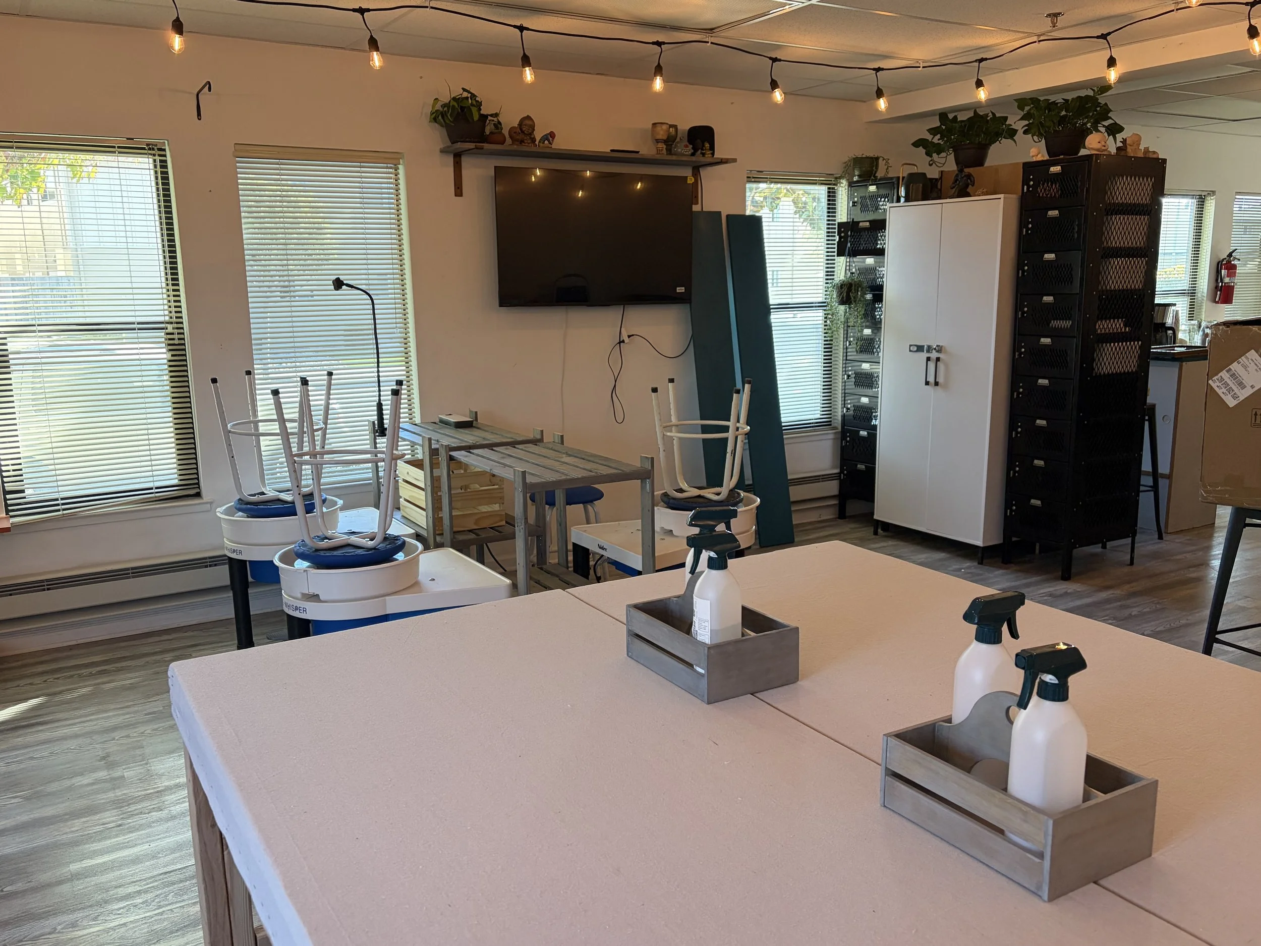 Room with large windows with blinds, a wall-mounted television, string lights hanging from the ceiling, plants and decorations on shelves, stackable chairs on upside down tables, a white cabinet, black storage racks, a cardboard box, and cleaning spr