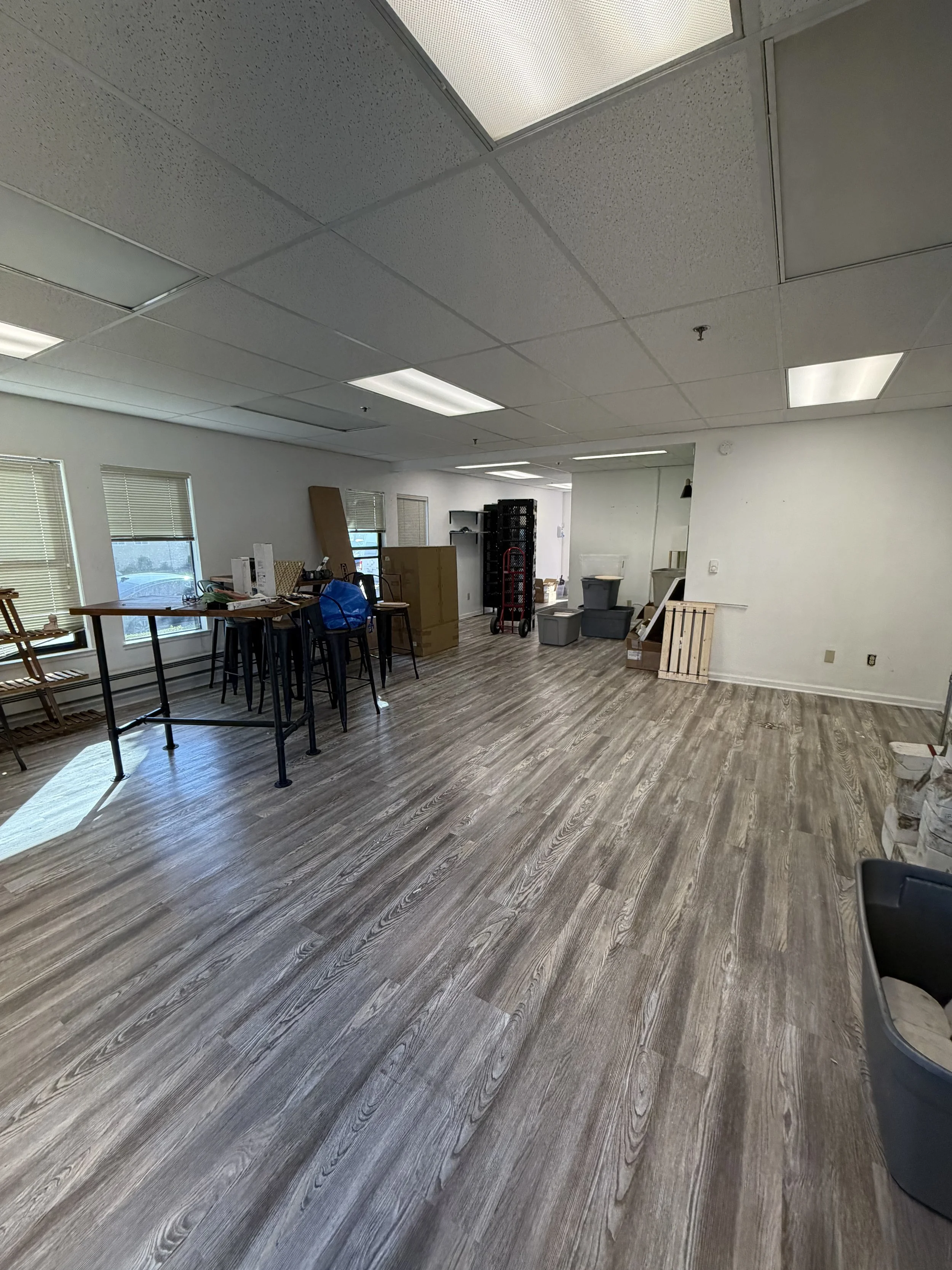 Empty room with wood flooring, some furniture and containers, sunlight coming through windows, ceiling lights, and a white wall.