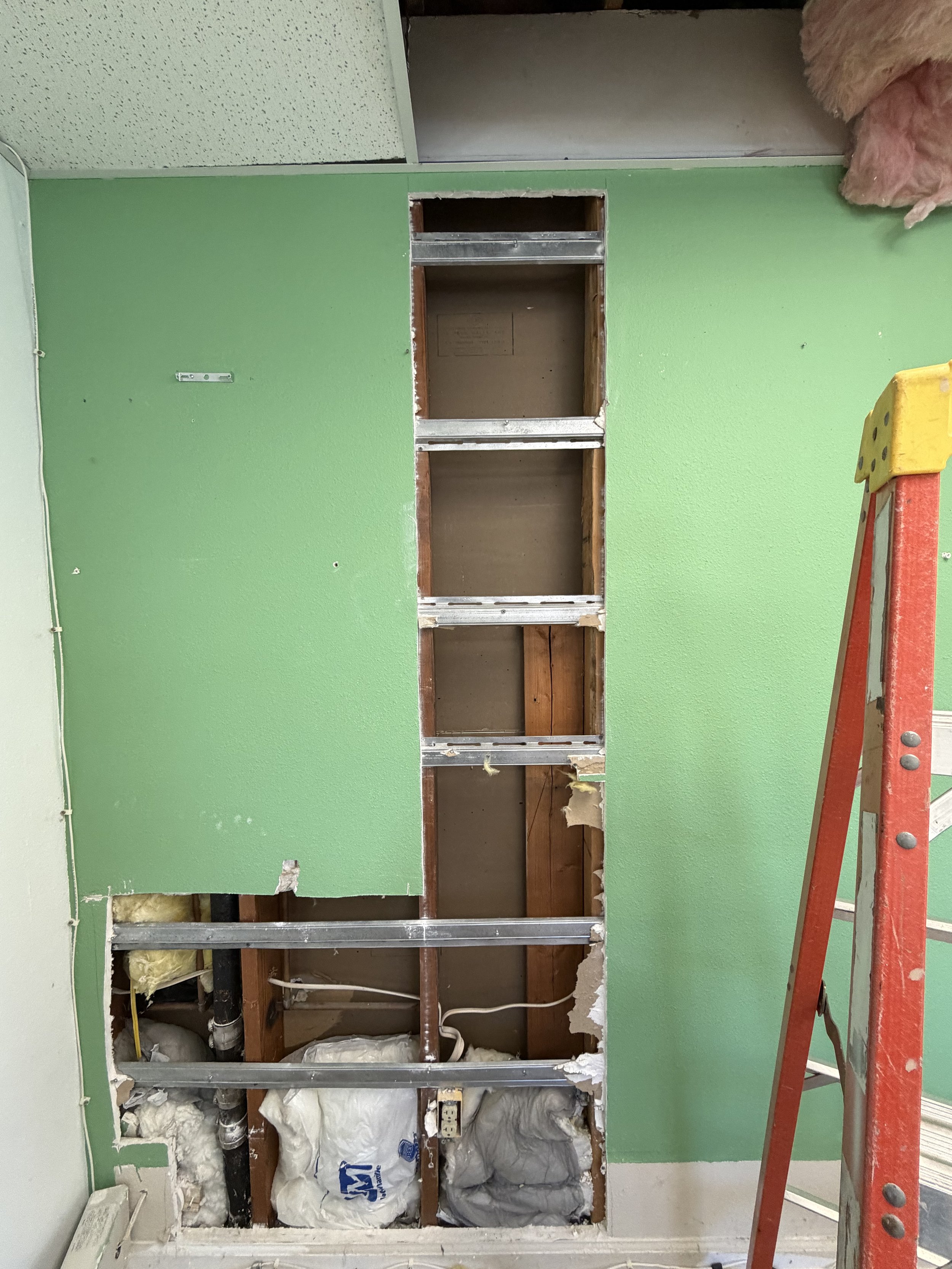 A wall with a large rectangular opening under renovation, showing exposed pipes, insulation, and wooden framing. The wall is painted bright green, and the surrounding areas show signs of construction work.