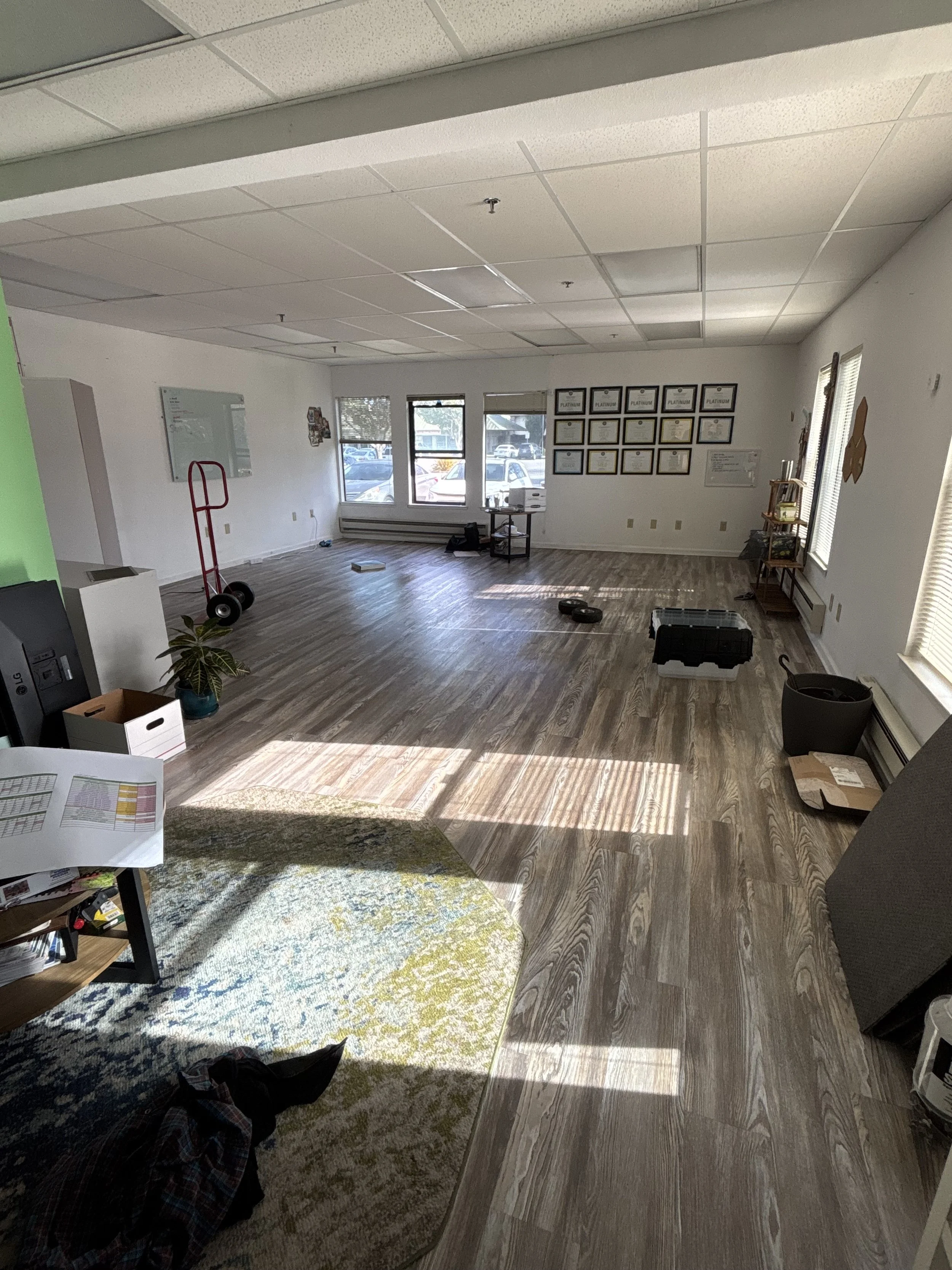 An empty office space with wooden flooring, white walls, windows allowing sunlight, framed awards or certificates on the wall, and various office supplies and equipment scattered around.