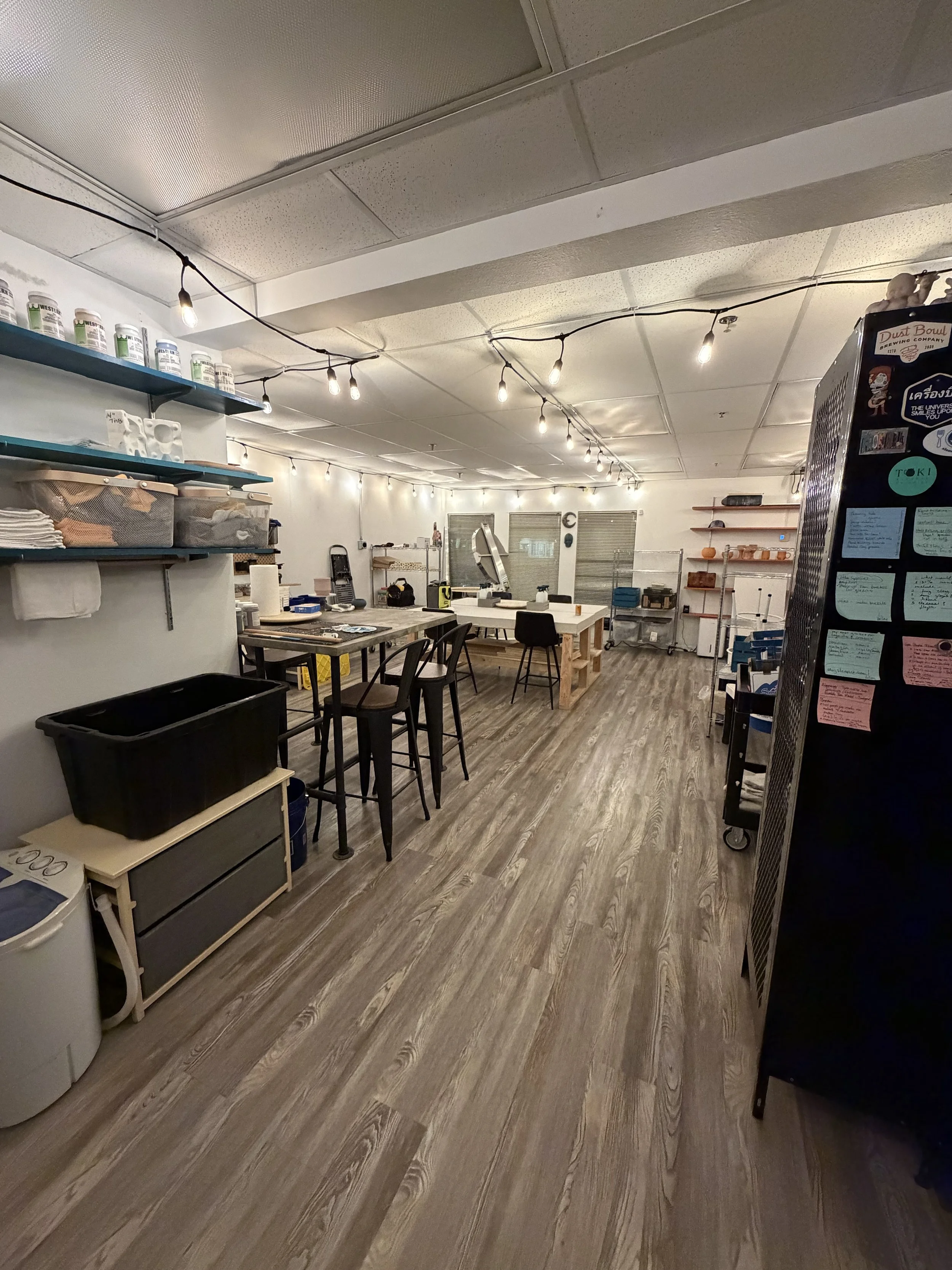 An industrial-style workspace with tables, chairs, shelves, storage bins, and string lights hanging from the ceiling.