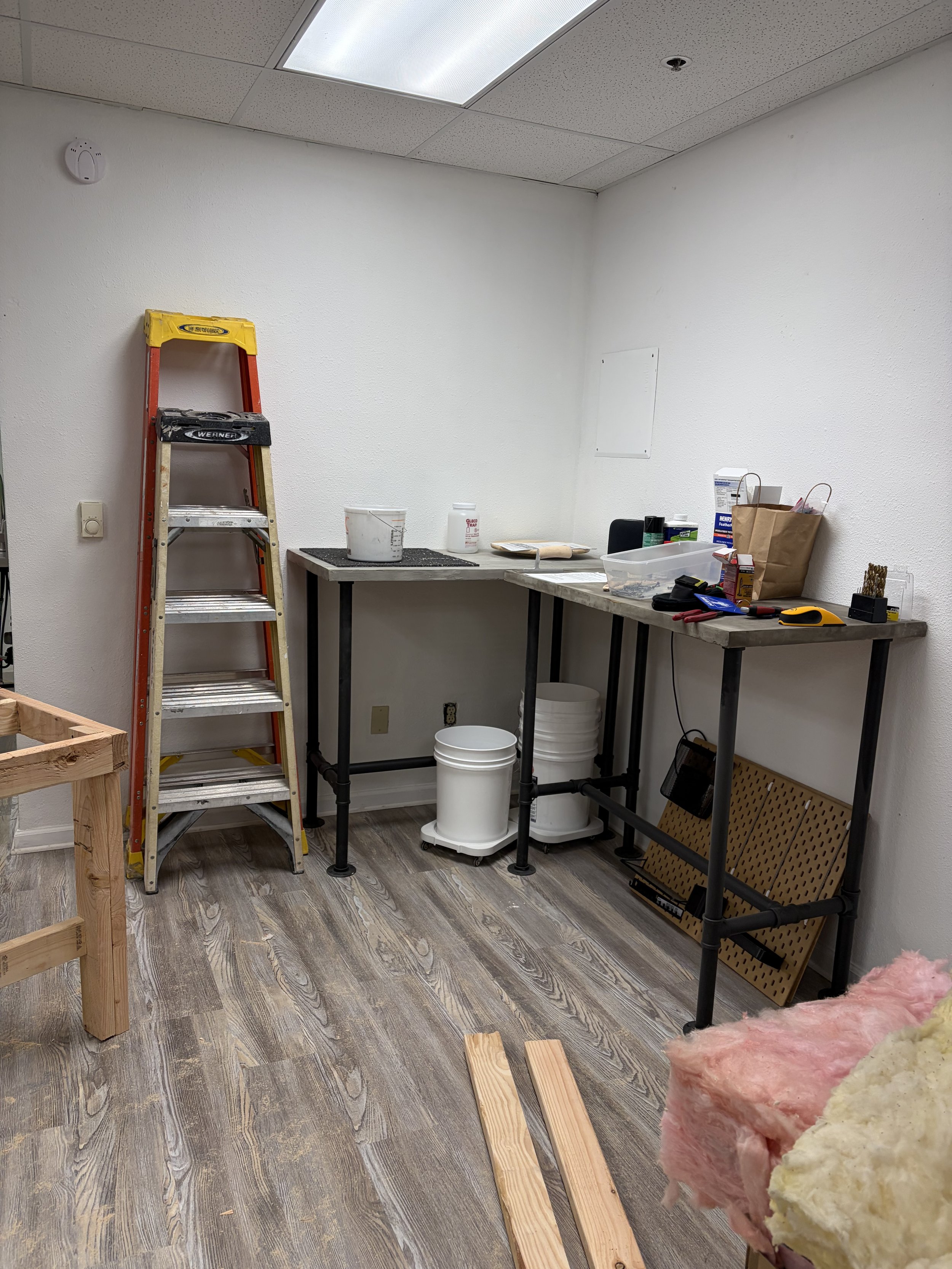 A room under construction with a yellow and red ladder, a work table with tools and supplies, and insulation in the foreground. The room has white walls, a drop ceiling with a light panel, and wood-look flooring.