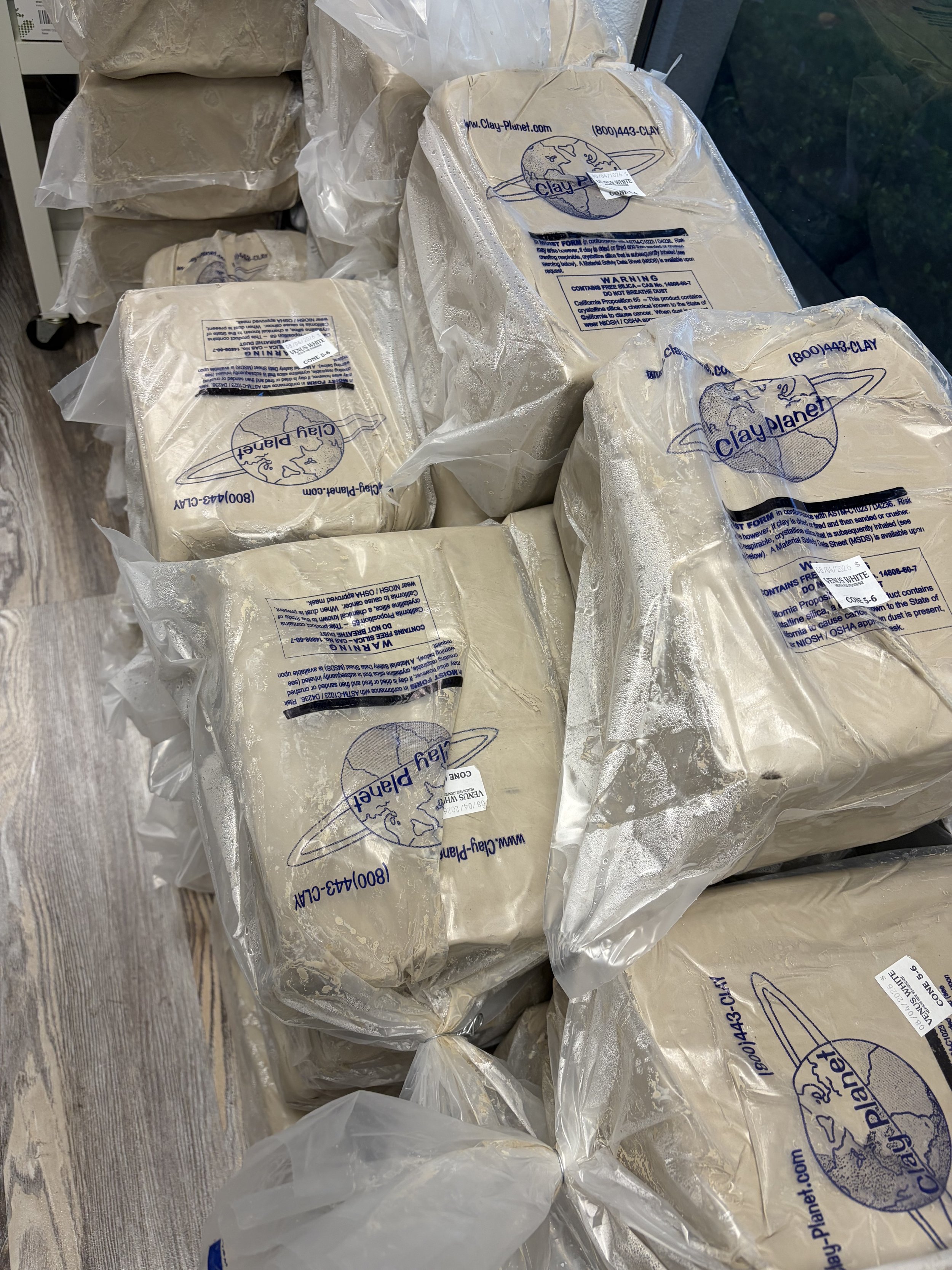 Several bags of white clay with labels, some open, on a wooden surface near a window.