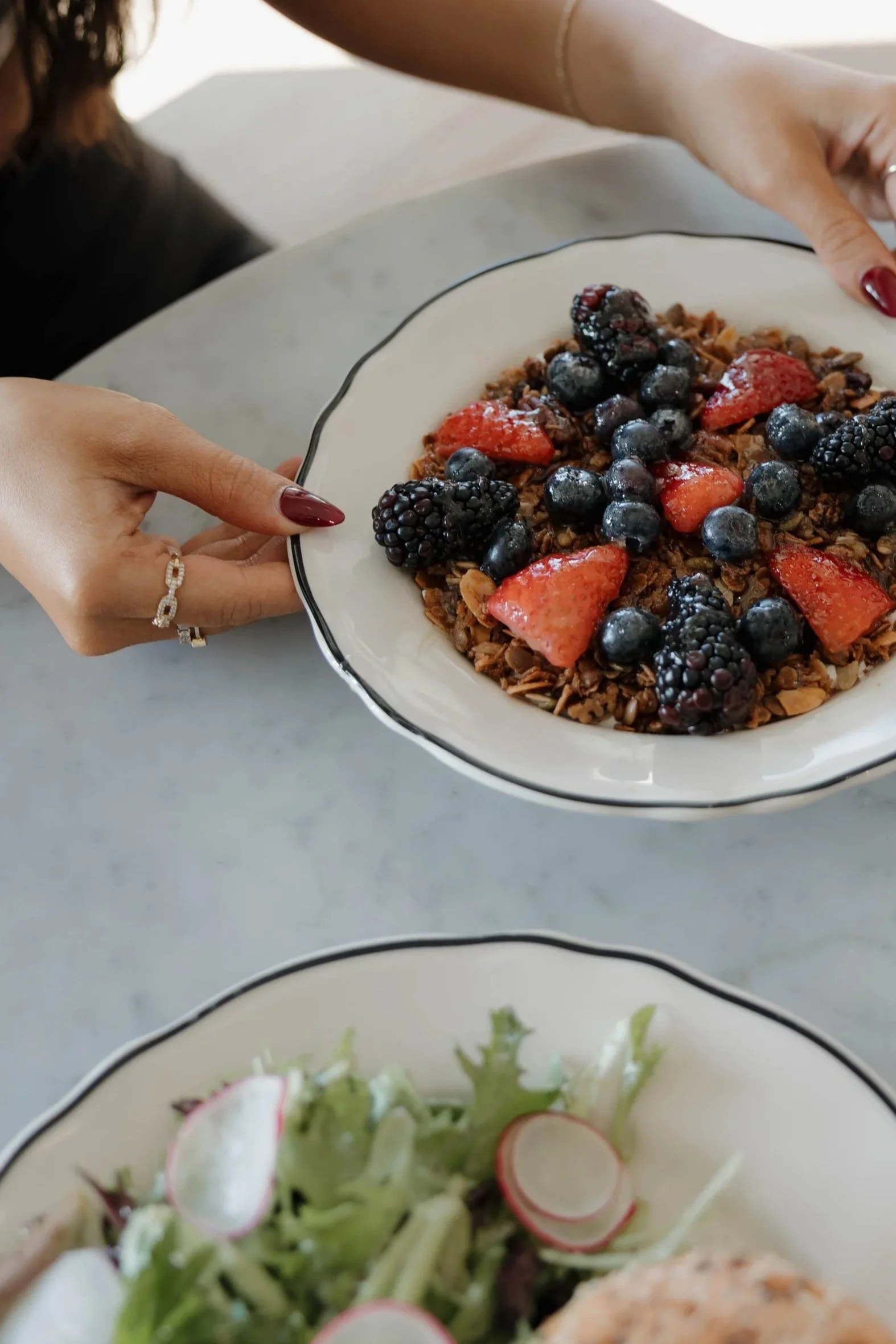 Person holding a bowl of mixed berries and granola at a table.