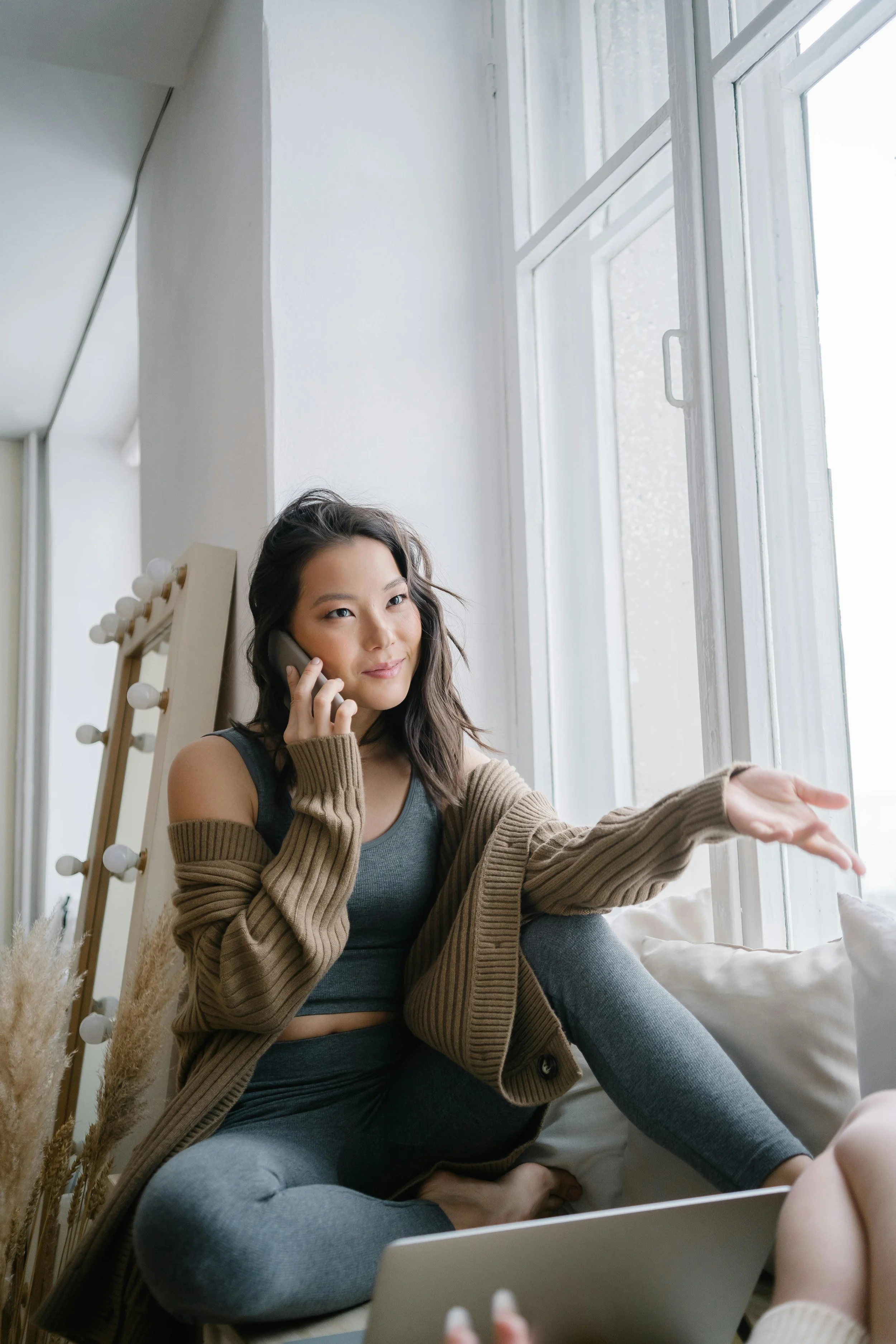 Young woman sitting on a couch, talking on the phone by a large window, gesturing with her hand.