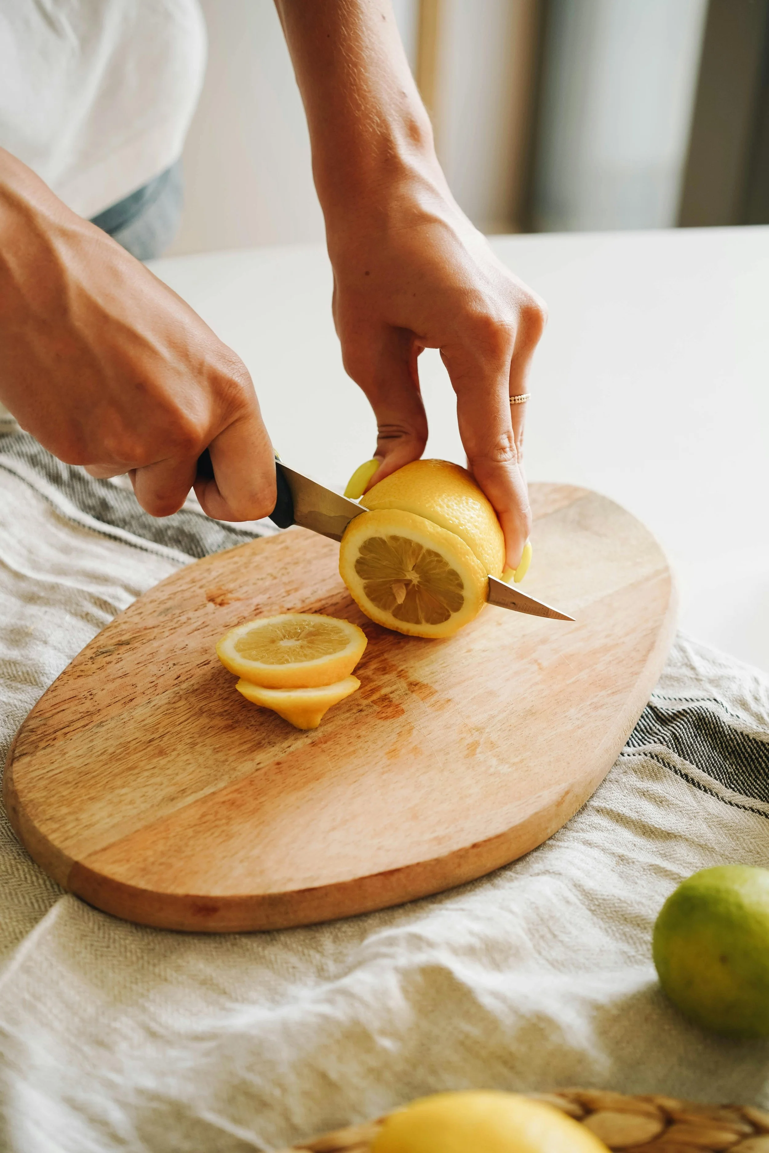 Person slicing a lemon on a wooden cutting board with a knife, with slices of lemon on the board and a whole lemon nearby.