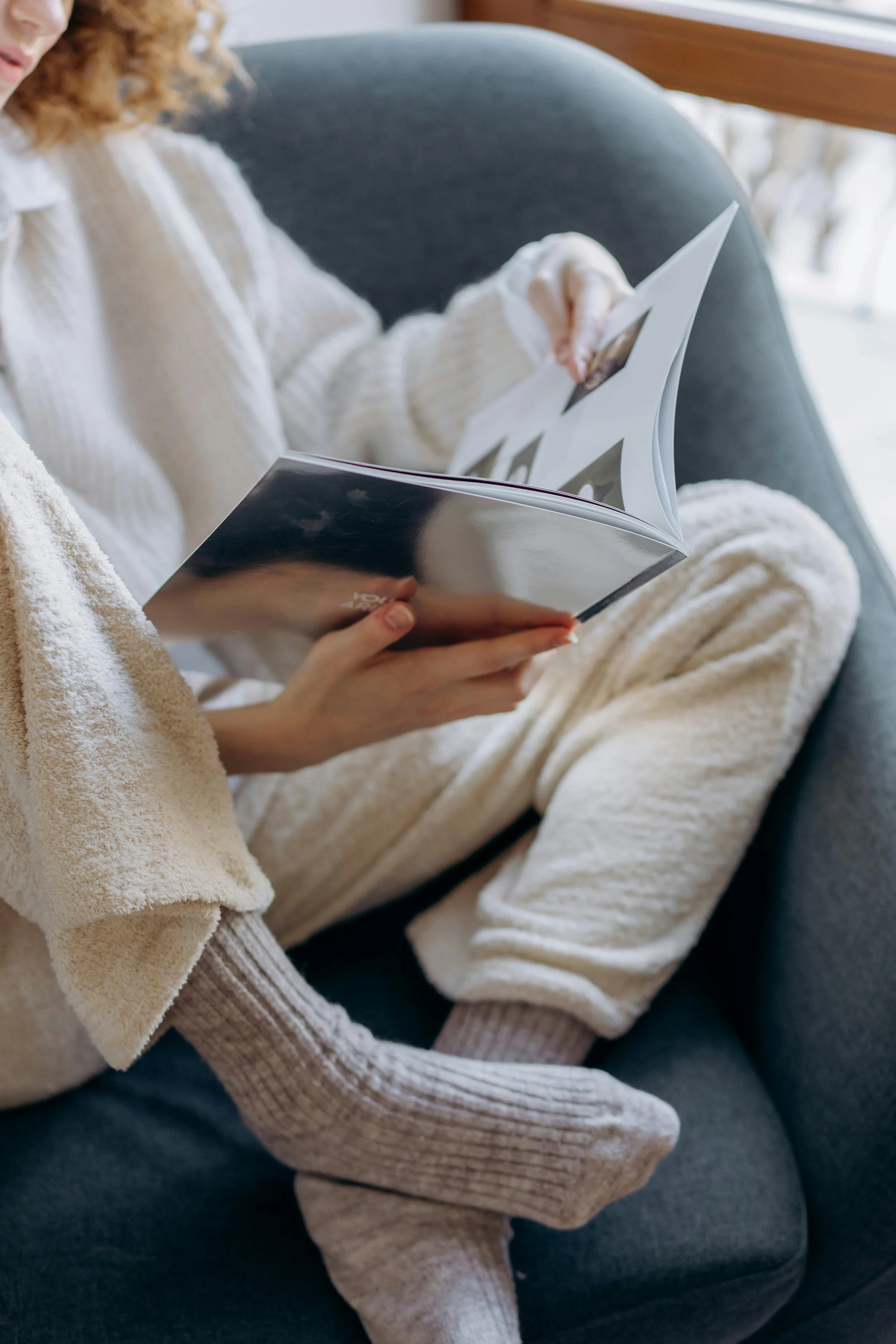 A woman in cozy loungewear sitting on a gray couch, reading a magazine.