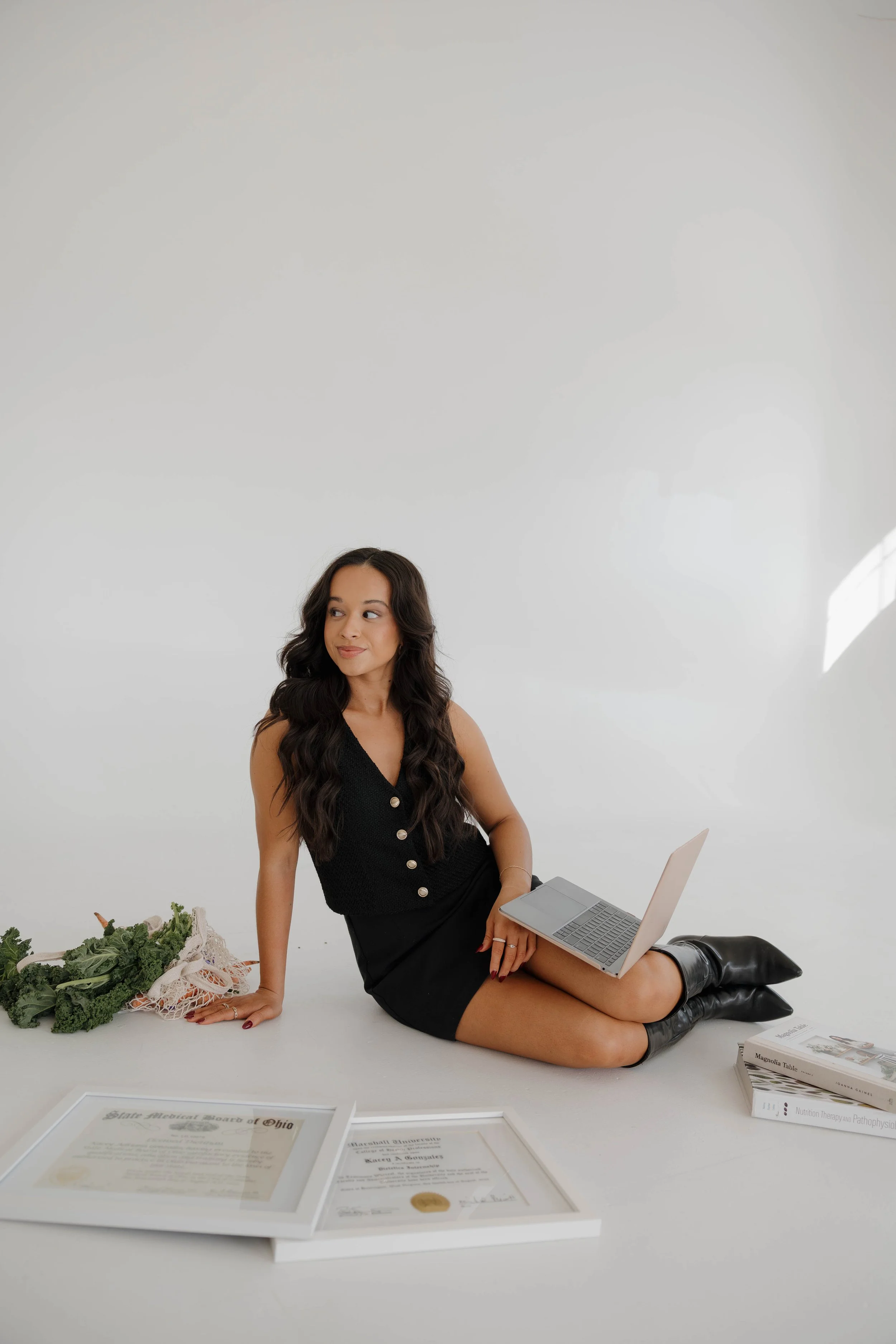 A woman with long dark curly hair sitting on the floor with a laptop on her lap, surrounded by some certification papers, a bunch of fresh kale, and a few magazines against a plain white background.