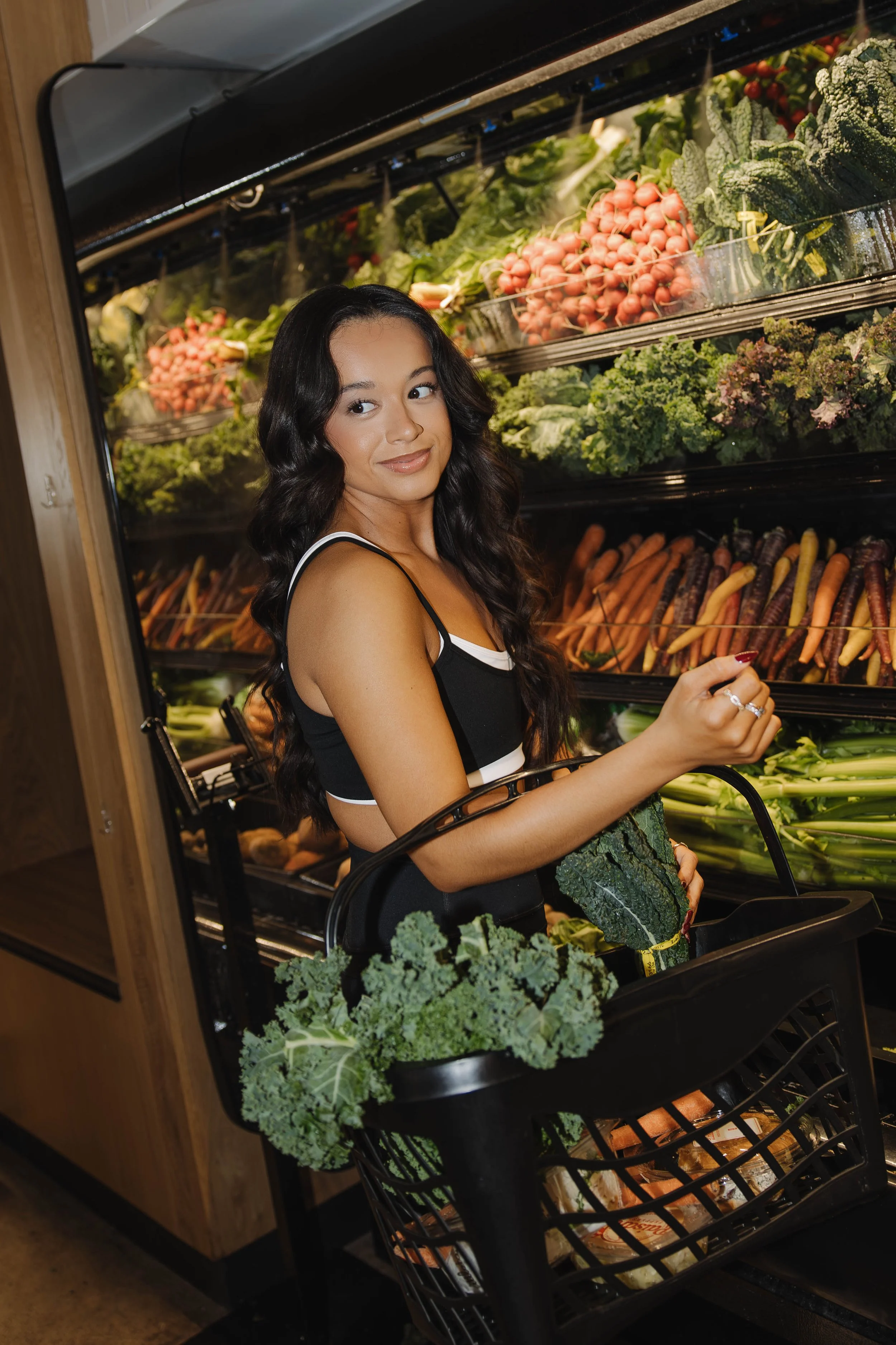 A woman grocery shopping, holding a cauliflower and a kale bunch, standing in front of a display of fresh vegetables at a grocery store.