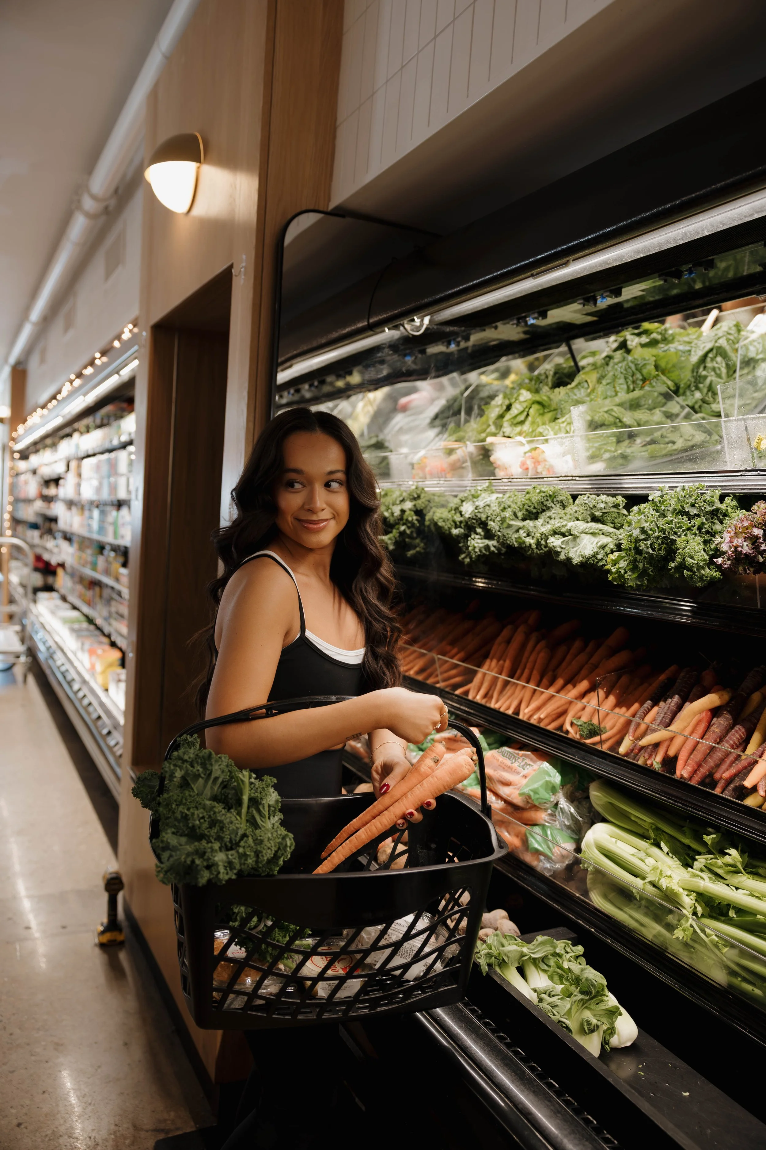 Woman shopping for fresh vegetables in a grocery store, holding carrots and a bunch of kale, standing next to a produce shelf filled with leafy greens and carrots.