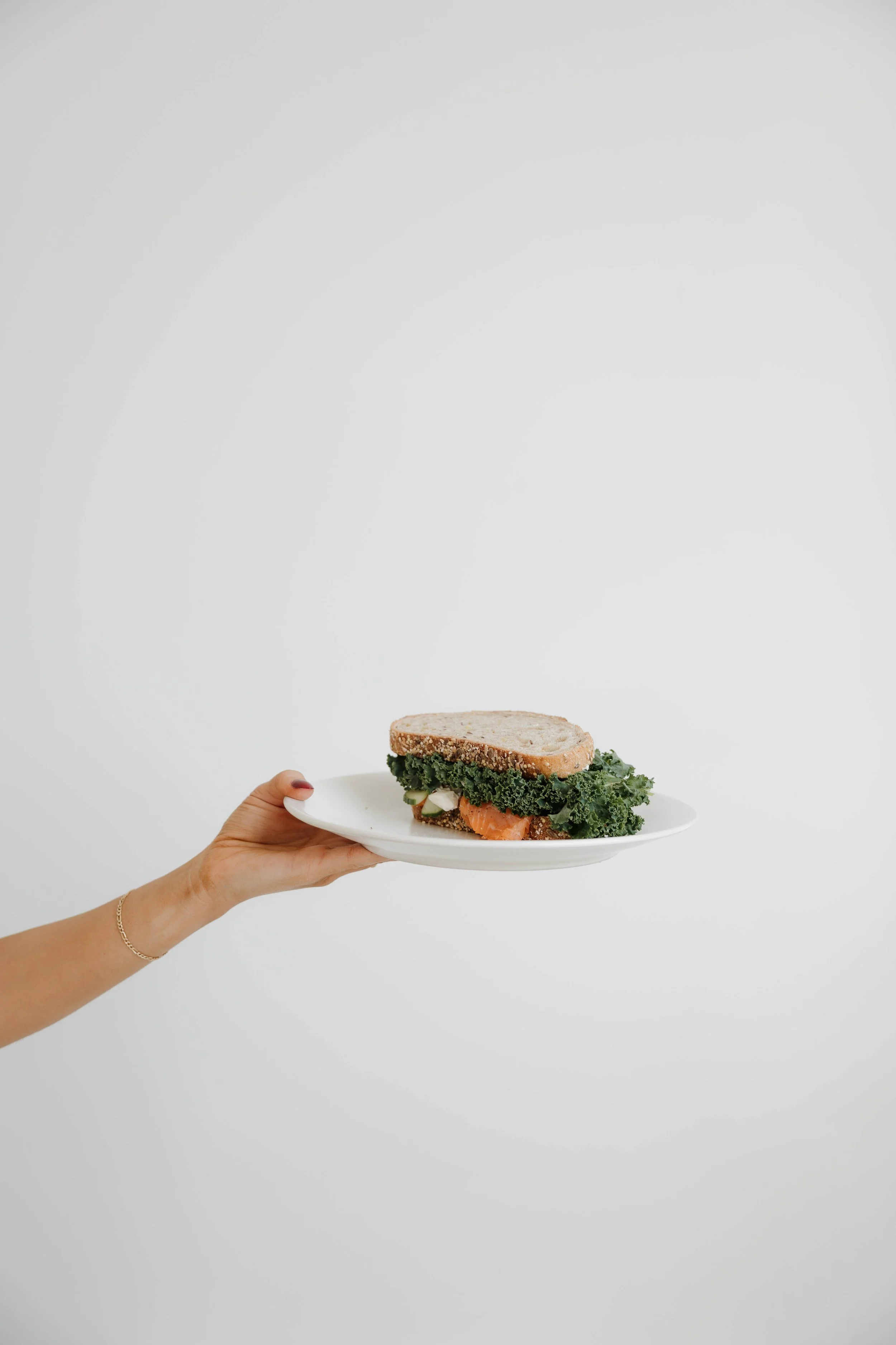 A hand holding a white plate with a sandwich containing kale, cucumber, and salmon against a plain white background.